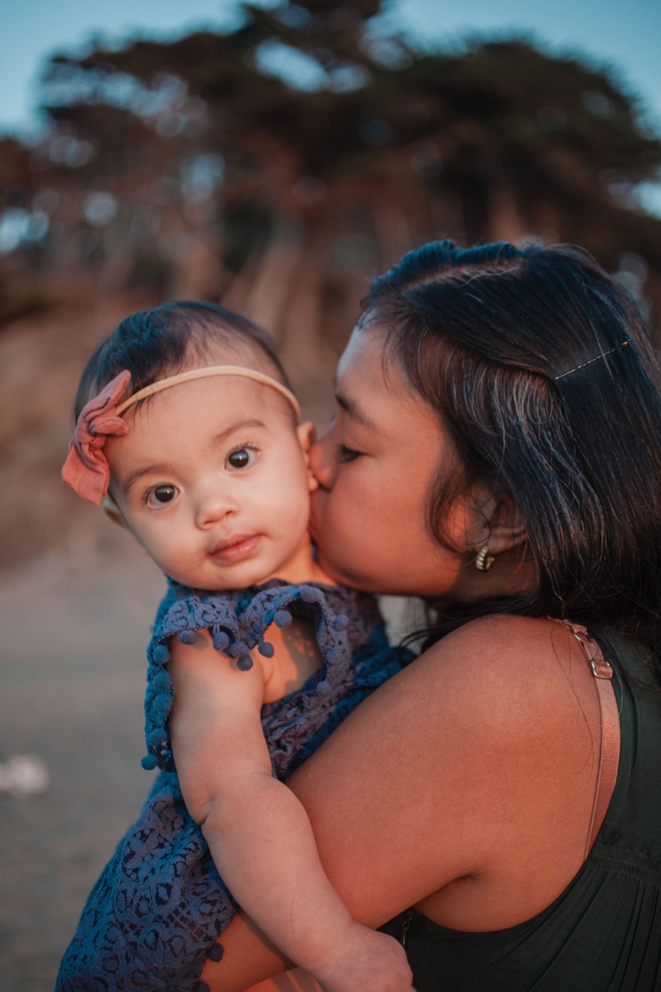 Mother kisses her infant daughter's cheek at golden hour, the toddler gazing directly at camera with wide eyes and serene expression