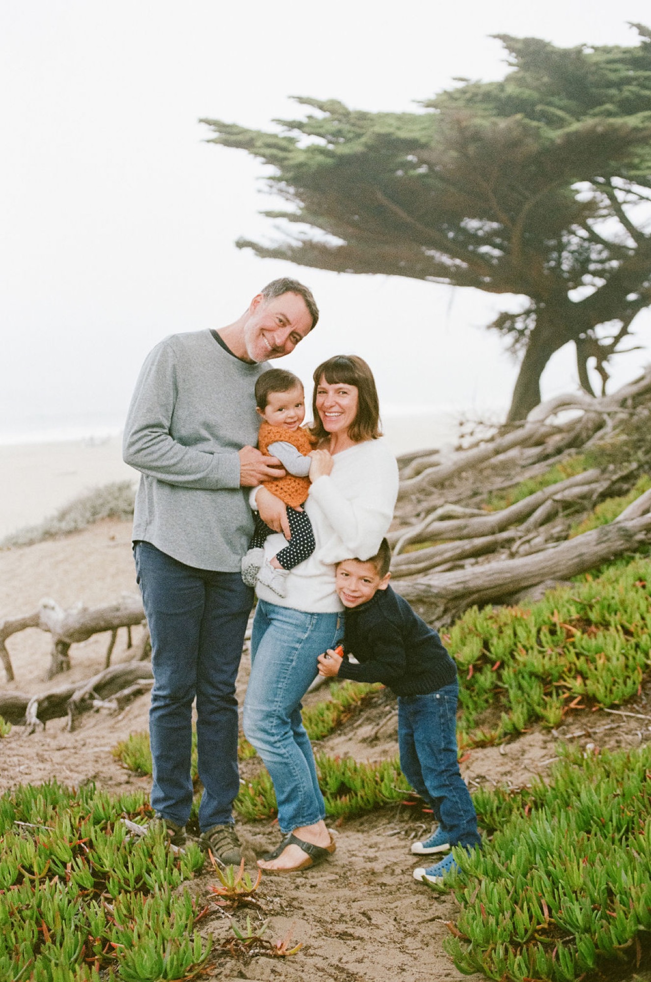 A family of three stands on a windswept coastal trail, the father smiling while holding their toddler between his and the mother's embrace