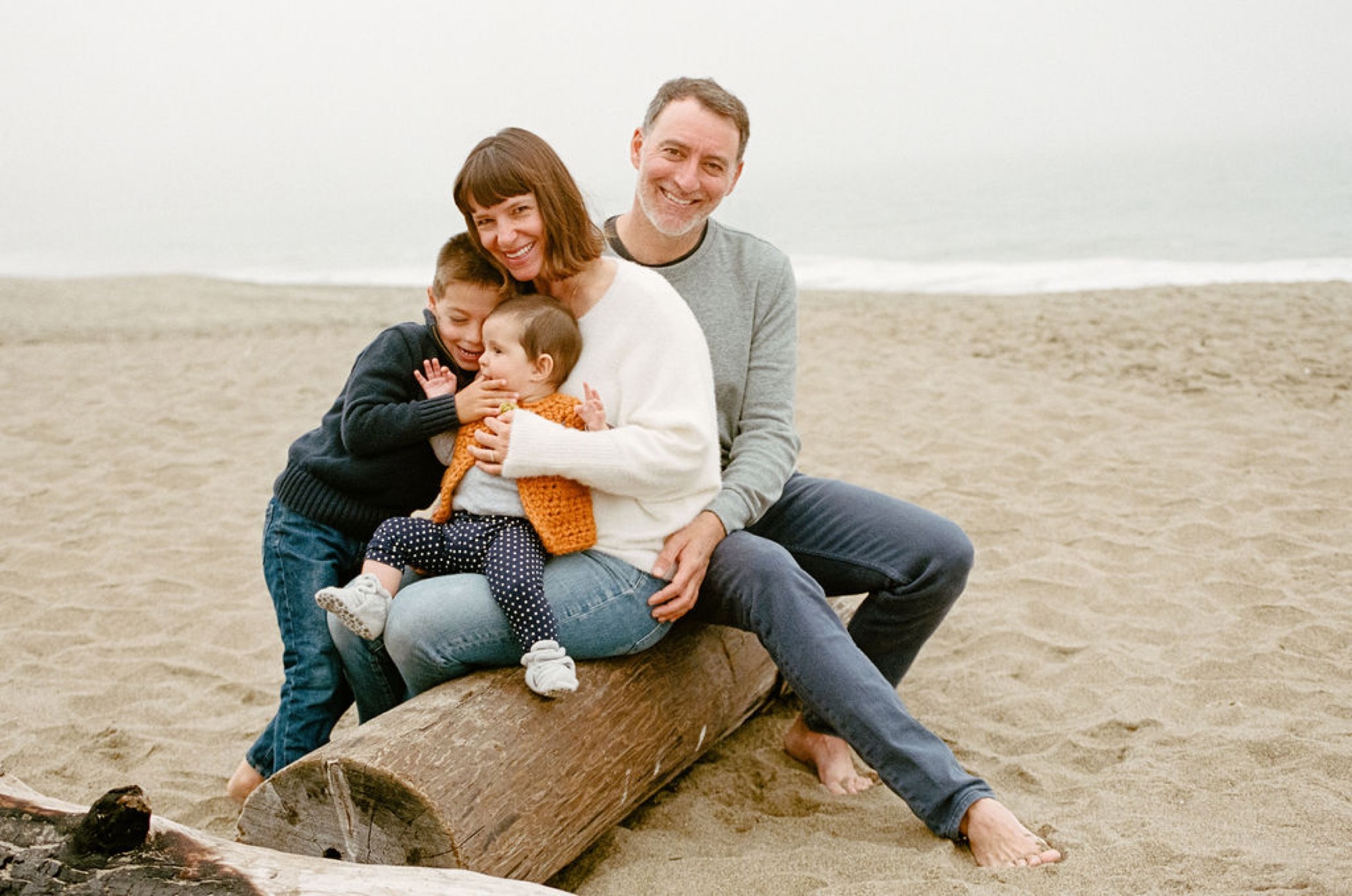 A family of four sits together on driftwood at the beach, with the father's arm around the mother and two young children nestled between them