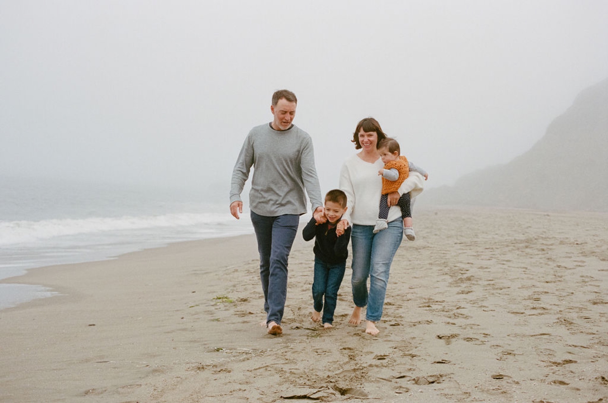 A family of four walks barefoot across misty Baker Beach, their relaxed strides and casual attire contrasting with the moody, fog-shrouded coastline behind them