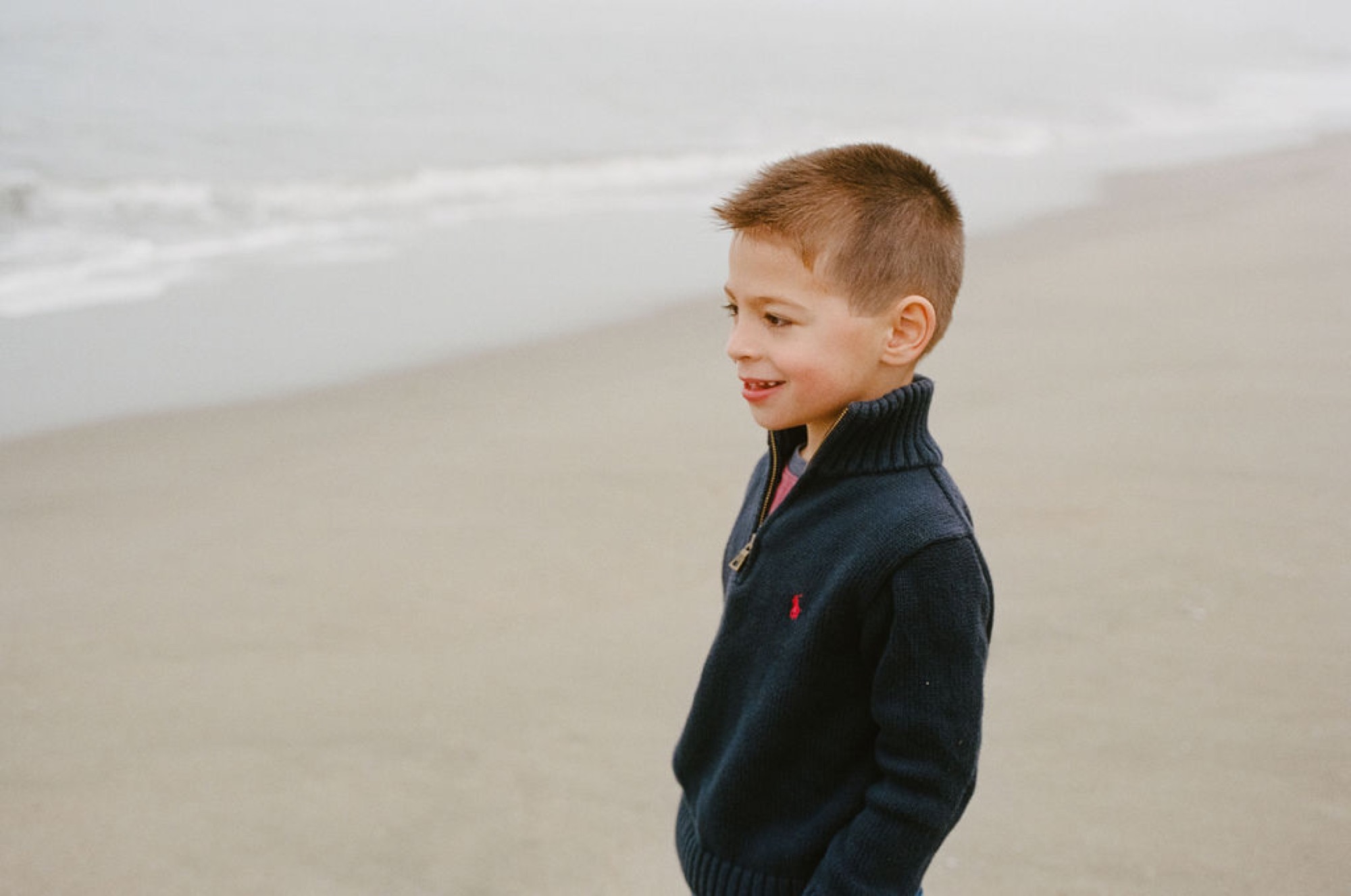 Young boy in navy jacket smiles while standing on misty beach sand, ocean waves visible behind him
