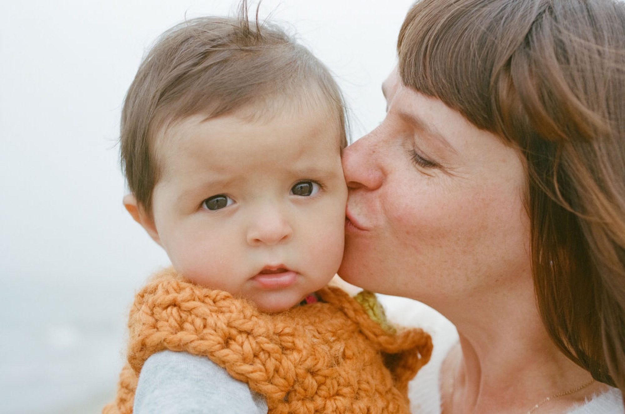 A mother kisses her infant's cheek while the baby gazes forward, wrapped in an orange knitted sweater against a soft white background