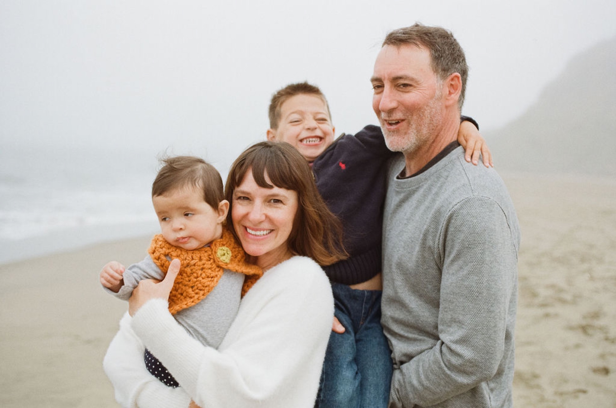 A smiling family of four stands together on a misty beach, with the mother holding a toddler in a golden sweater while the father and older son embrace them from behind