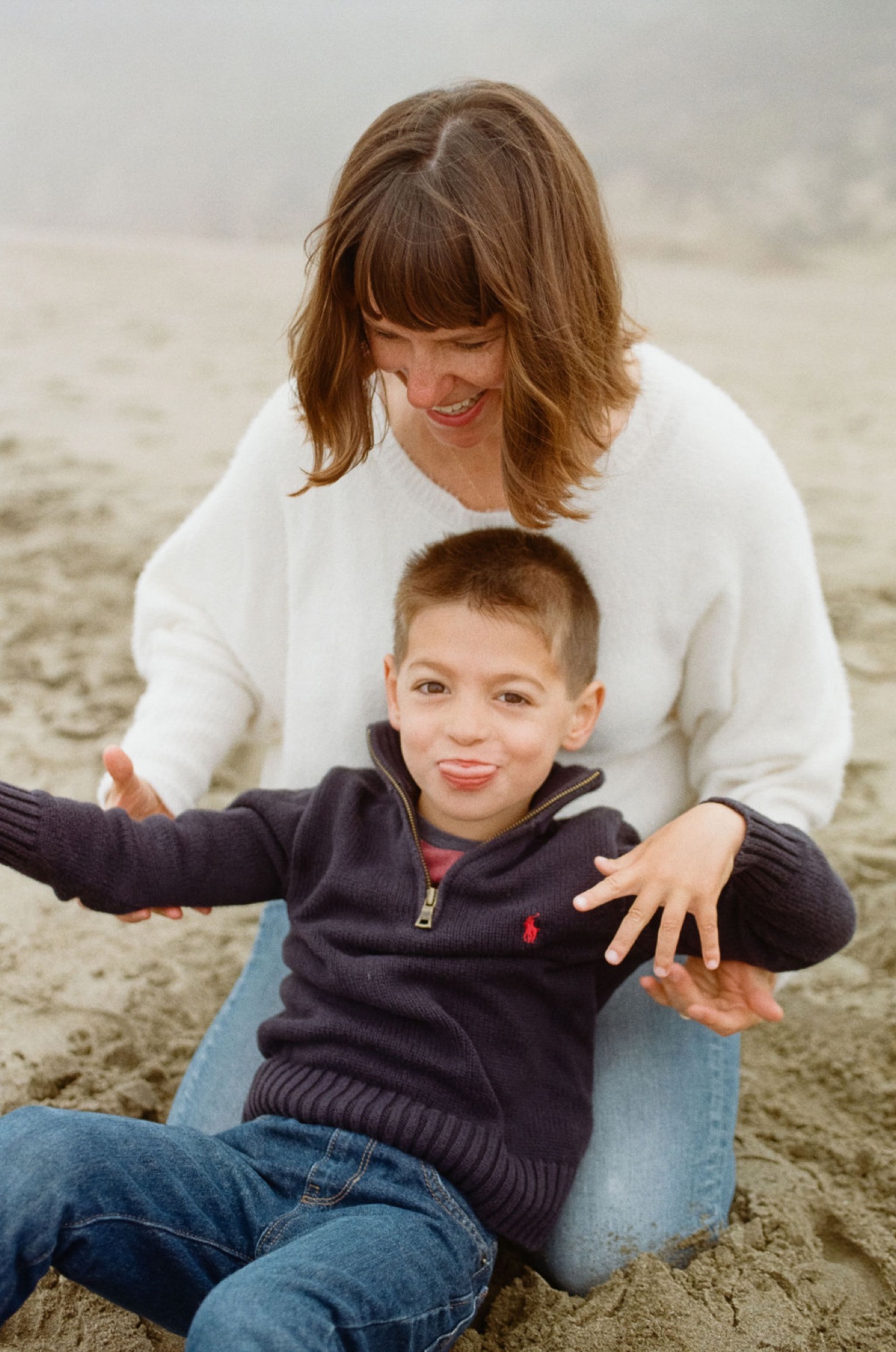 Woman crouches behind young boy on sandy beach, both smiling at camera with soft natural light