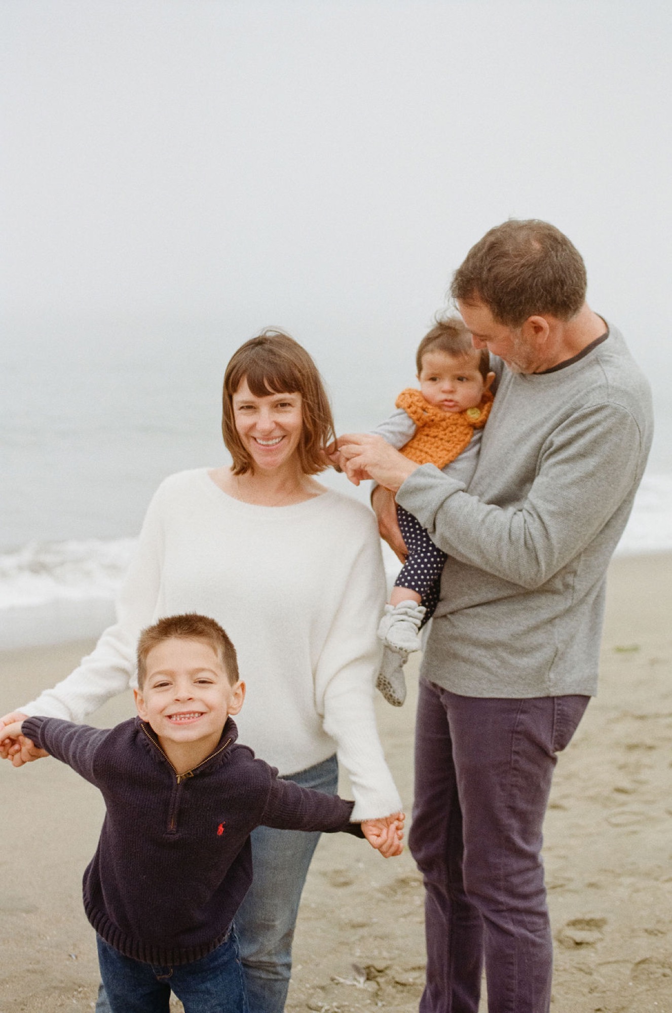 Family of four stands on misty beach holding hands, youngest child's arms spread wide with genuine joy and spontaneity