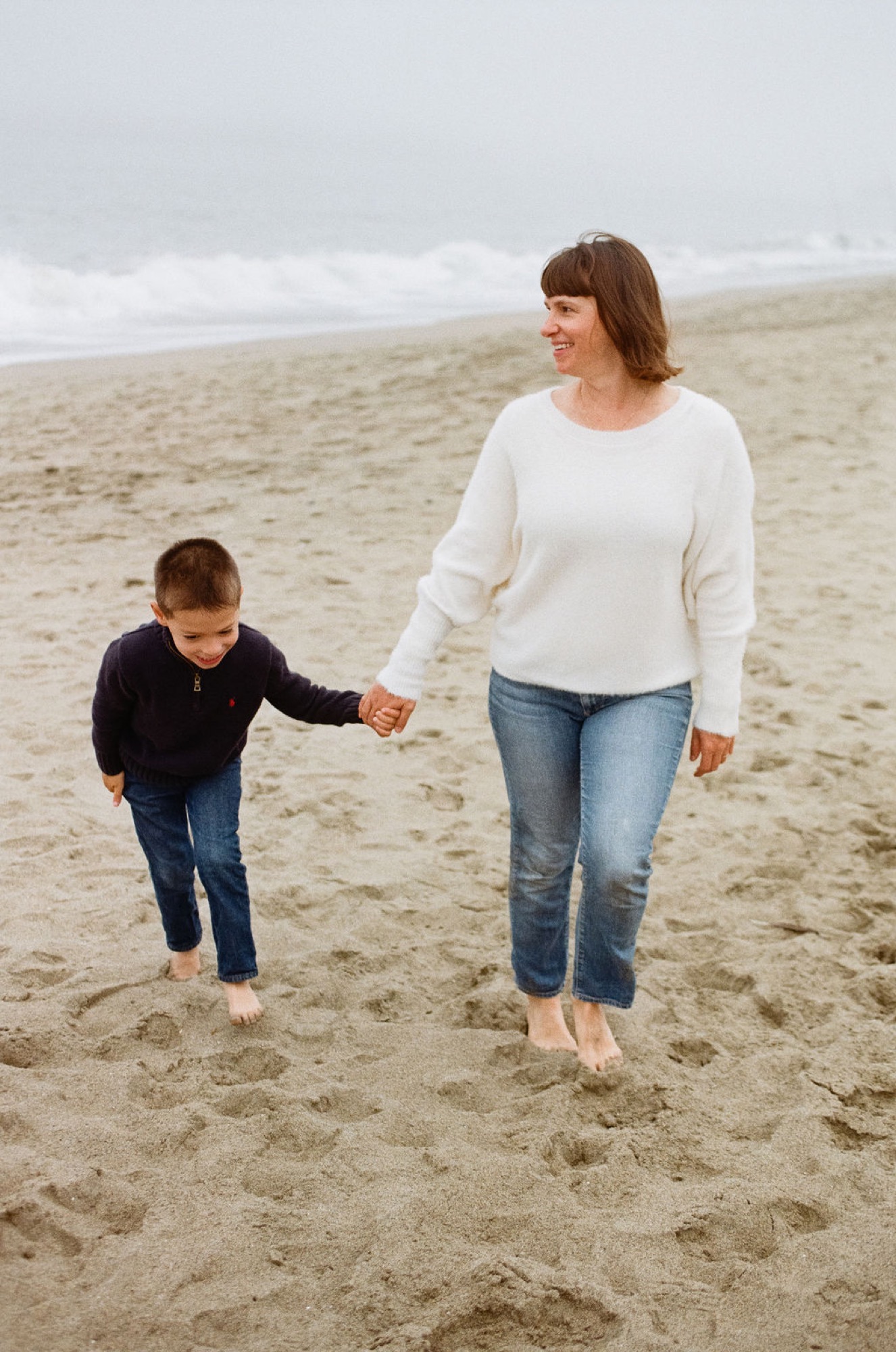 Woman and young boy walk barefoot across wet sand, holding hands, with overcast sky and waves behind them