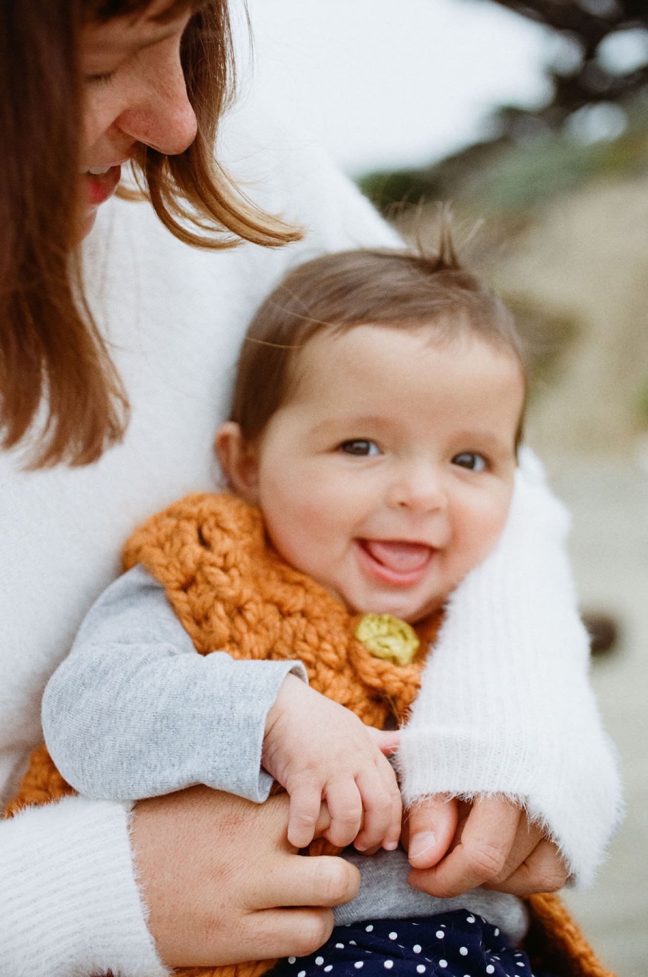 A beaming toddler in a golden knit sweater grins widely while held by a smiling parent against a soft, blurred coastal backdrop