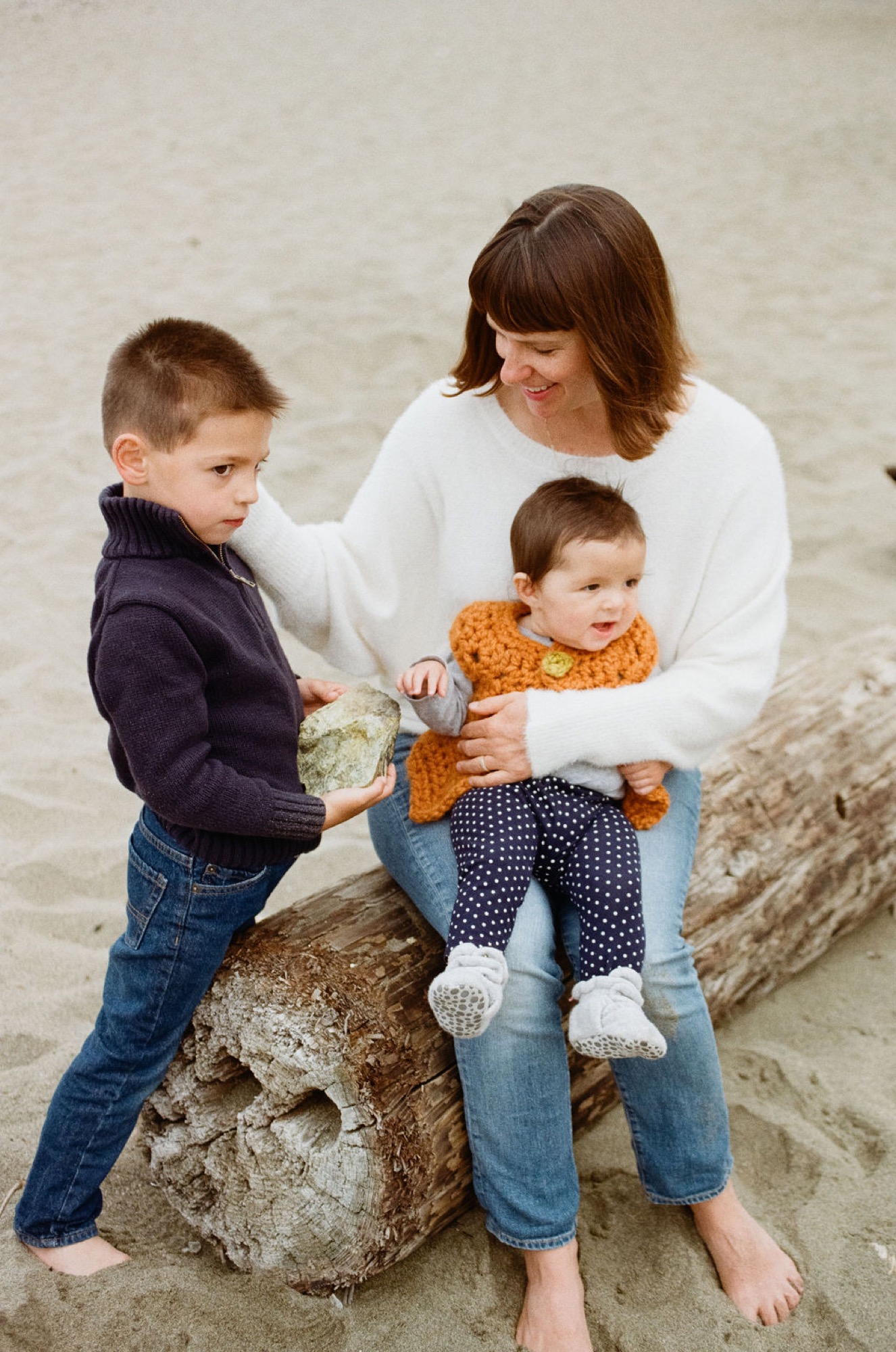 Woman and two young boys sit on a weathered driftwood log at the beach, the older child holding a shell while the younger one clutches an orange toy