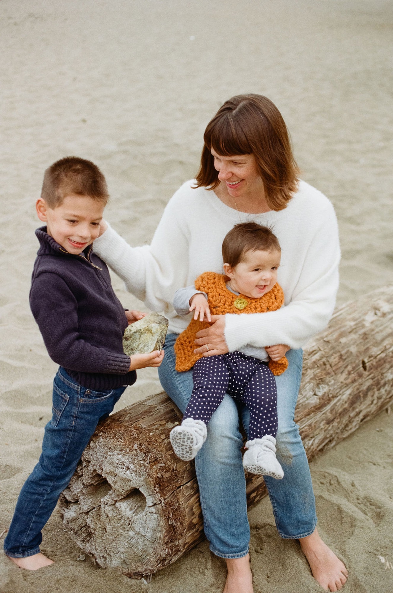 A mother sits on a weathered beach rock with her two young sons, all smiling warmly at the camera in soft natural light