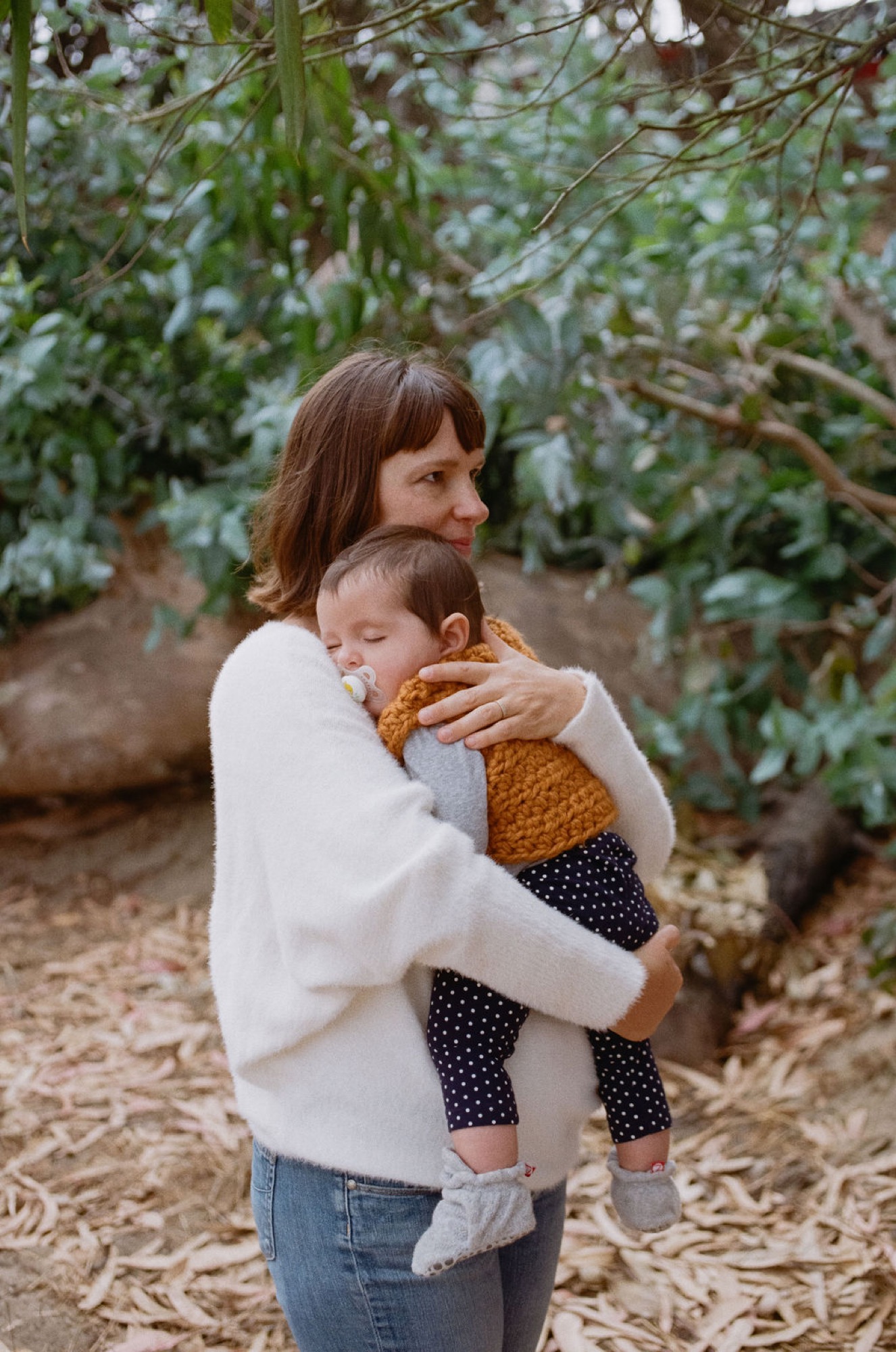 Mother holds toddler in golden-hour light among coastal scrub, child gazing downward in soft, natural embrace