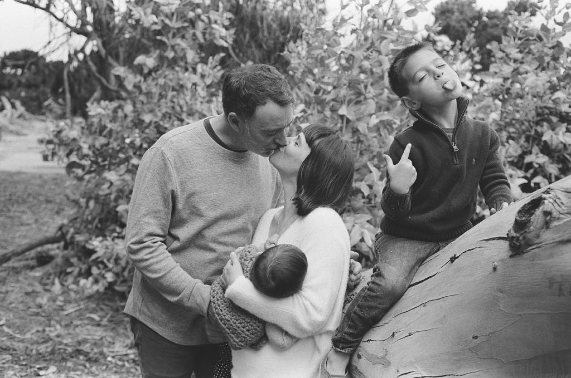 Father adjusts young child's clothing while mother holds infant, all gathered near gnarled tree trunk in dappled forest light