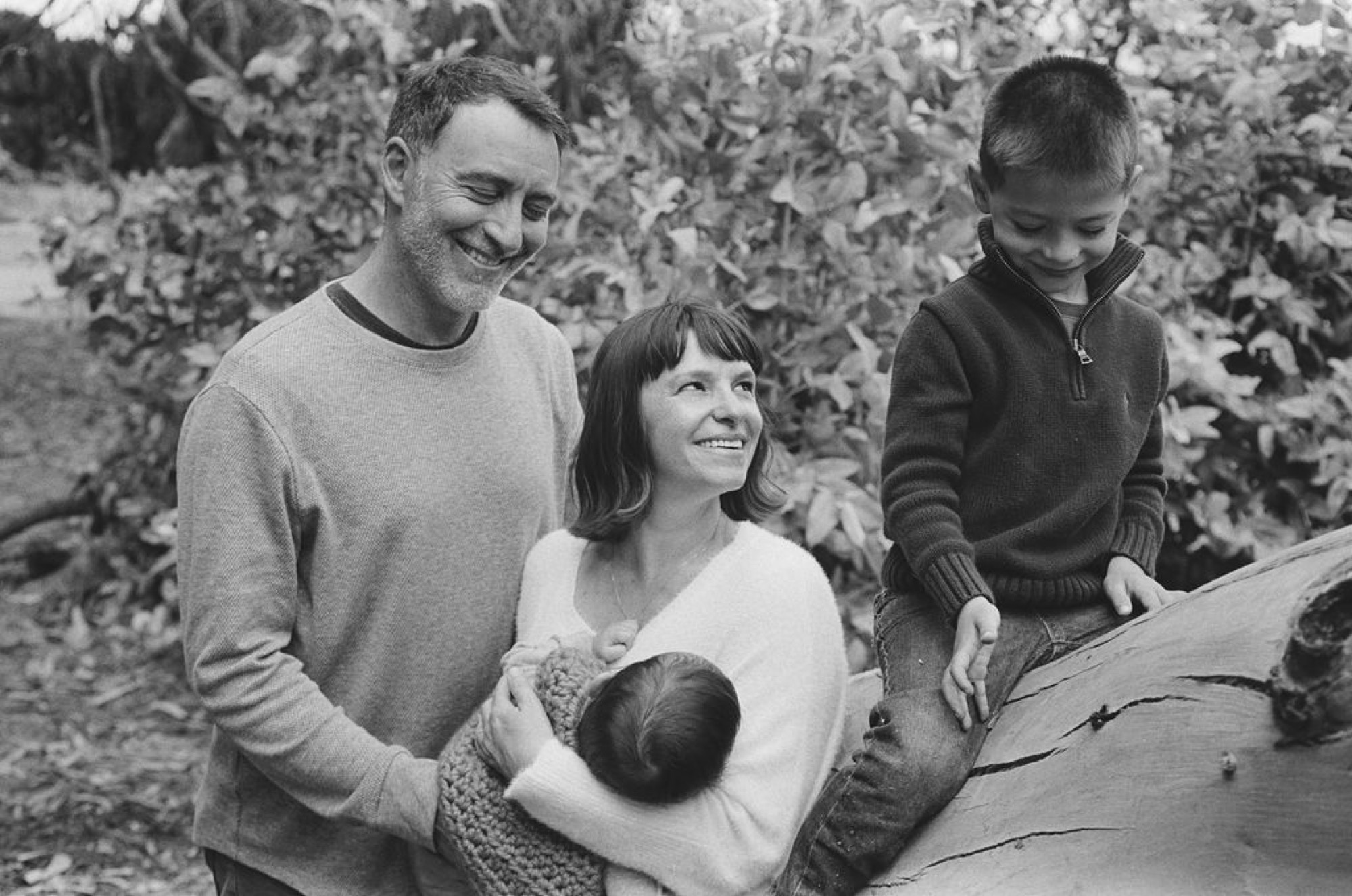 A family of four stands together among leafy vines, the parents flanking two children with genuine smiles in soft natural light