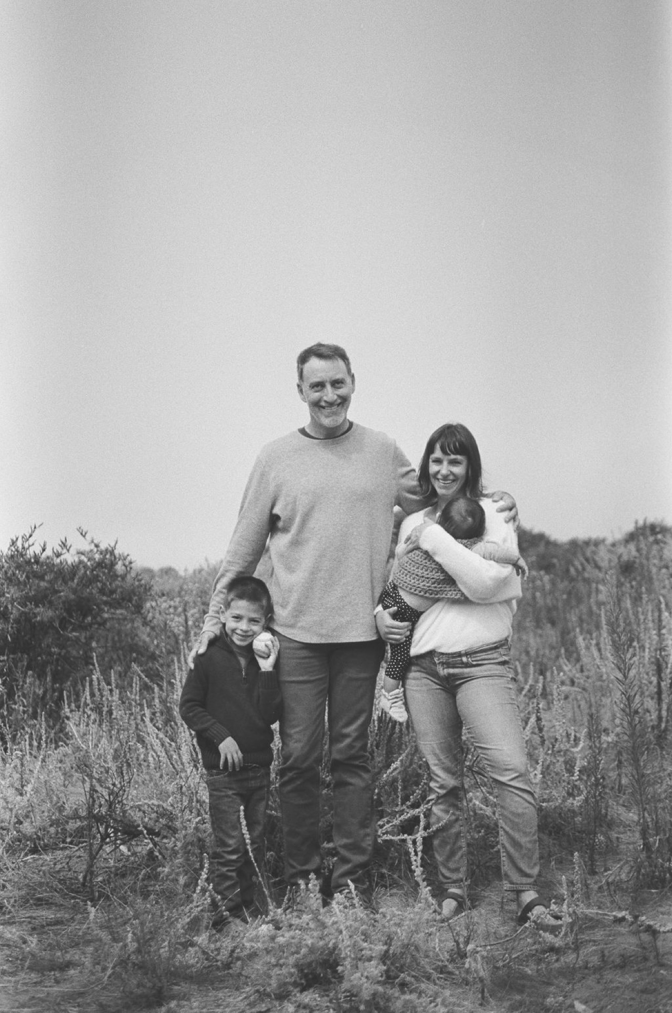 A man stands with three children in a grassy hillside field, all smiling warmly at the camera under soft natural light