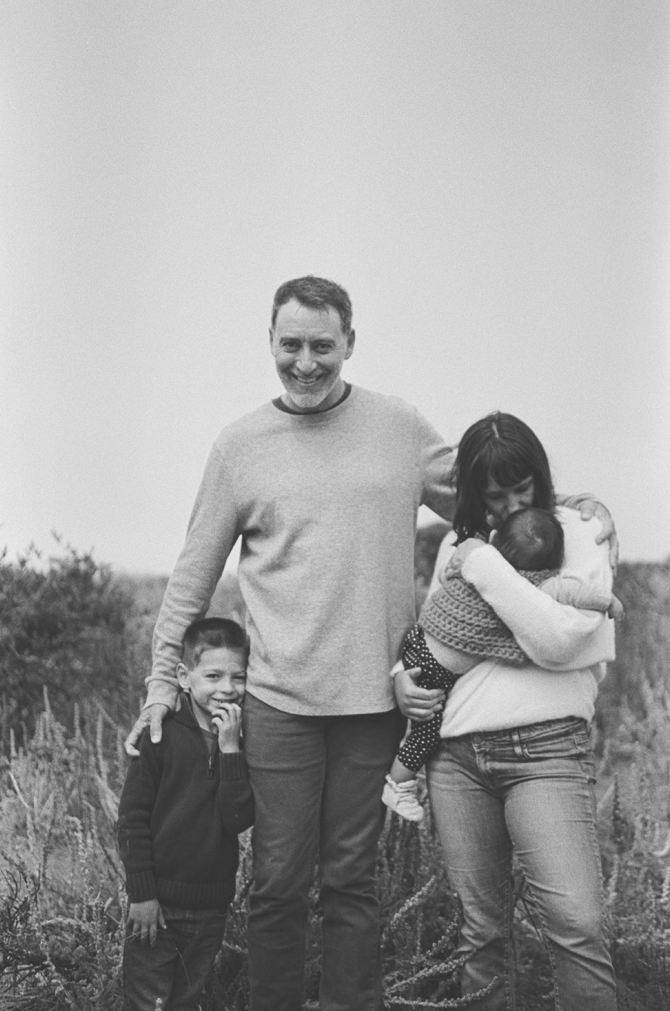 A man and two children stand together in an open grassy field, the smaller child nestled against his side while the older child leans in close