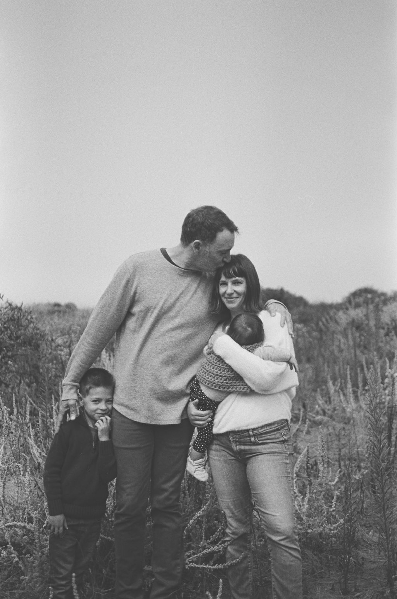 A father leans affectionately toward his wife while their young children stand close beside them in an open field, captured in warm natural light