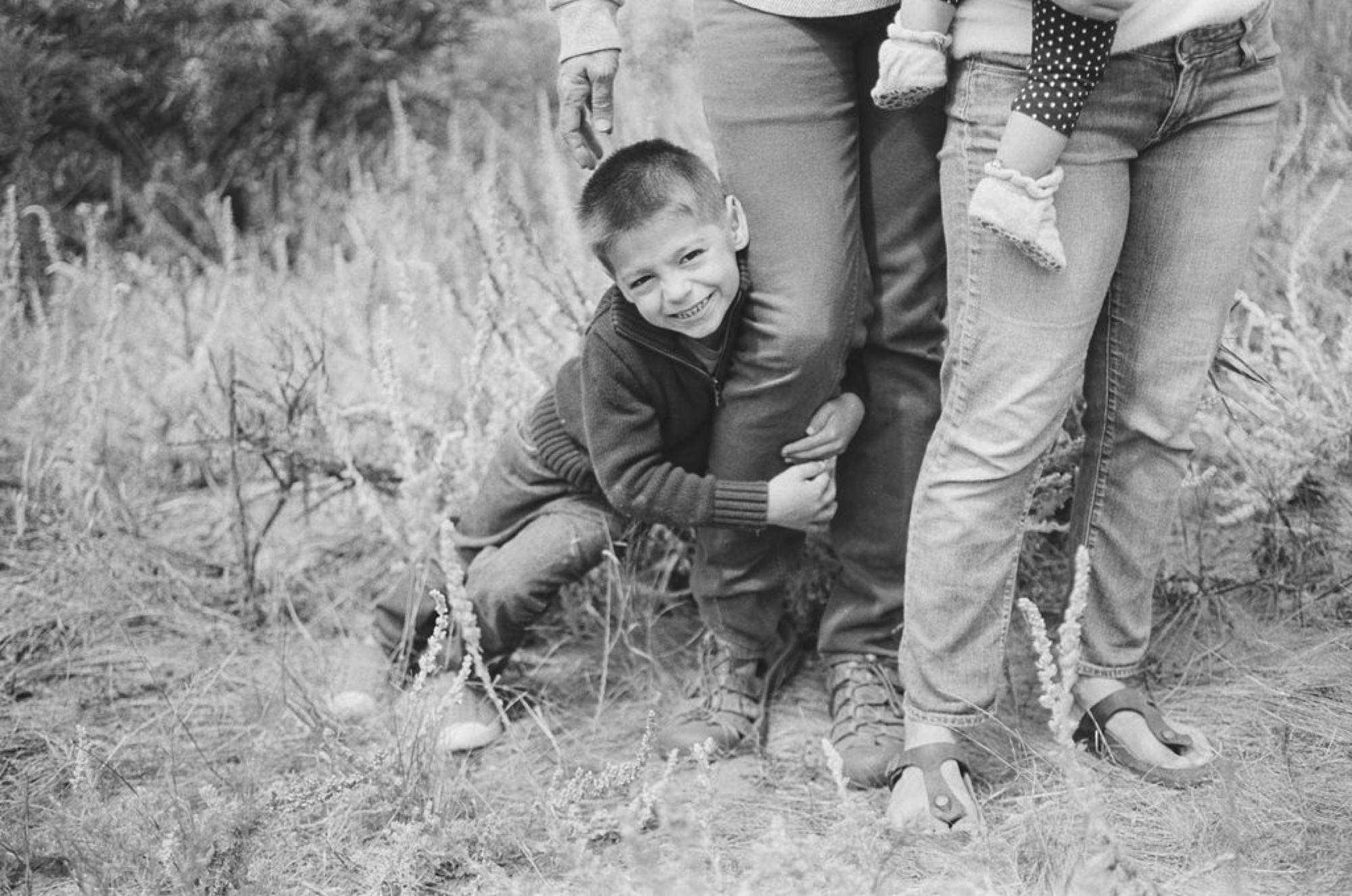 Young boy crouches beside adults' legs in dried grass, beaming with genuine delight at the camera