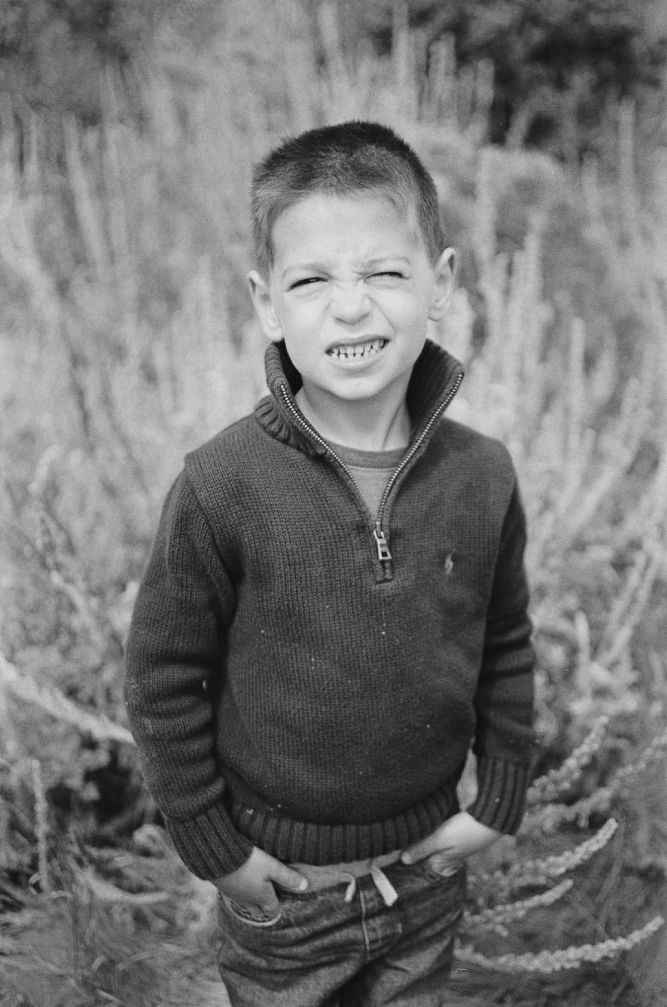 Young boy smiling broadly at camera, wearing dark quarter-zip sweater, standing amid dried coastal brush in natural afternoon light