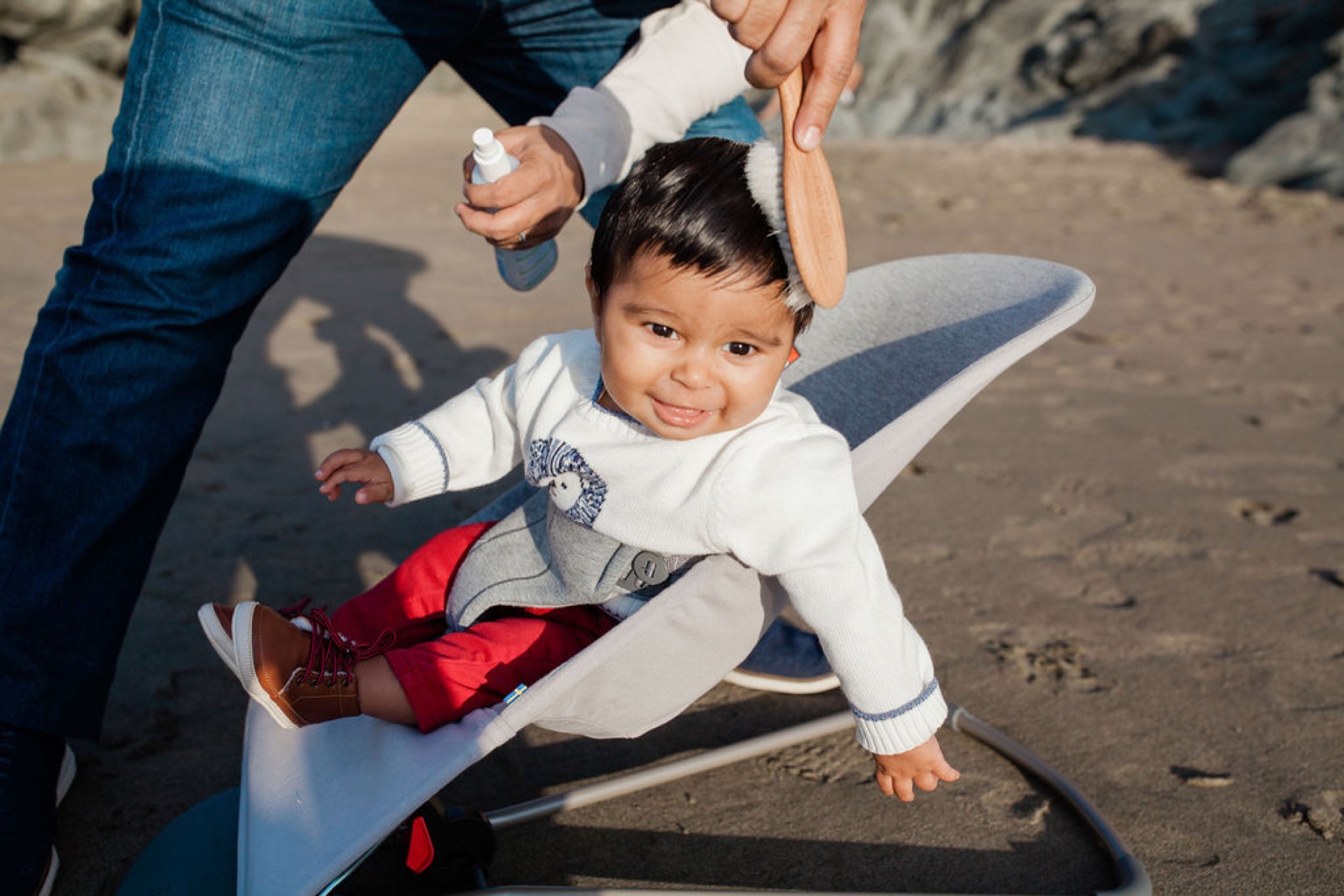 A toddler grins while being guided across beach sand by parents holding both hands, bright sunlight casting shadows