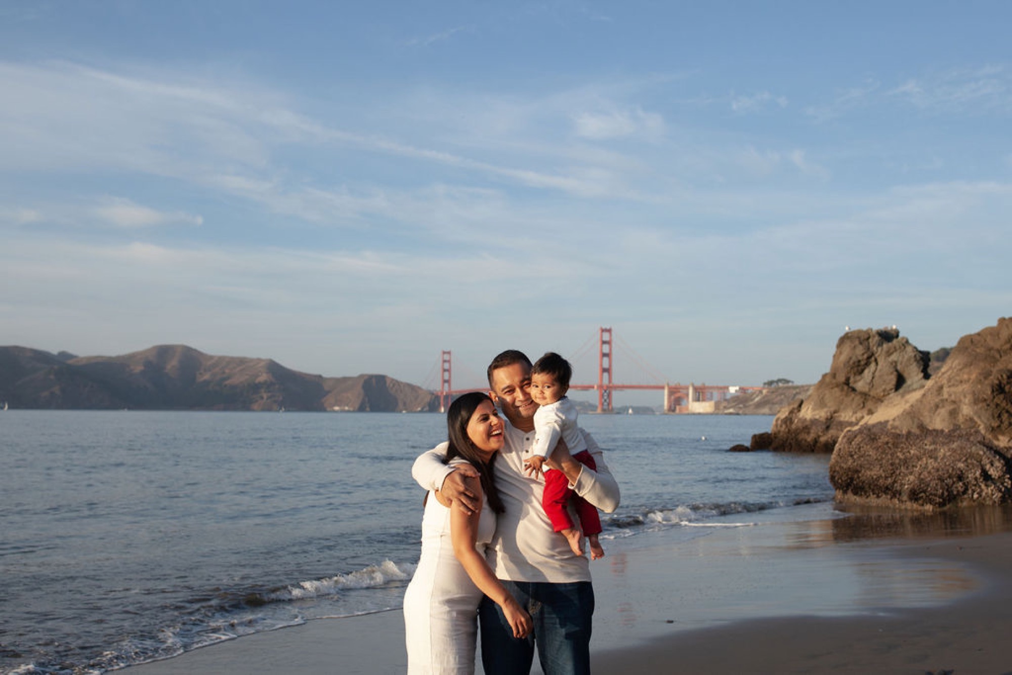 A man holds his young child while standing beside a woman on the beach, the Golden Gate Bridge gleaming red across the bay behind them