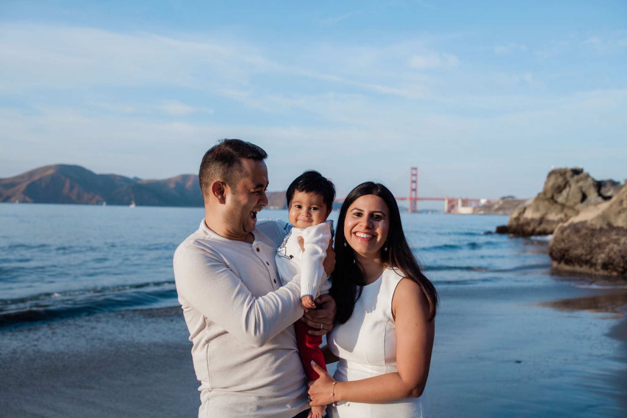 Family of three in coordinating white outfits smile genuinely at the camera while standing in shallow surf, with the Golden Gate Bridge visible across the bay
