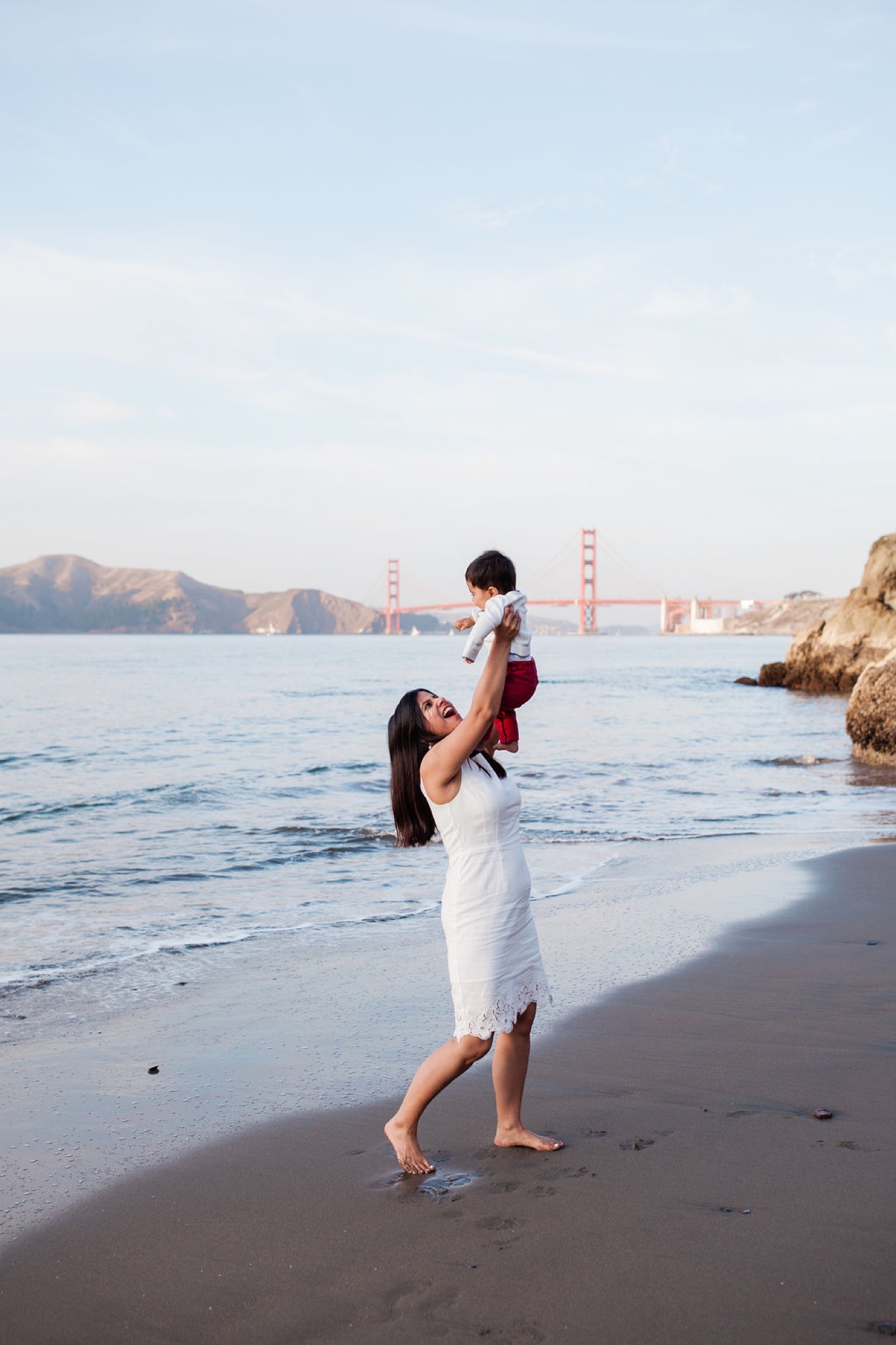 A mother lifts her young child skyward on the beach, both laughing joyfully with the Golden Gate Bridge glowing in the distance