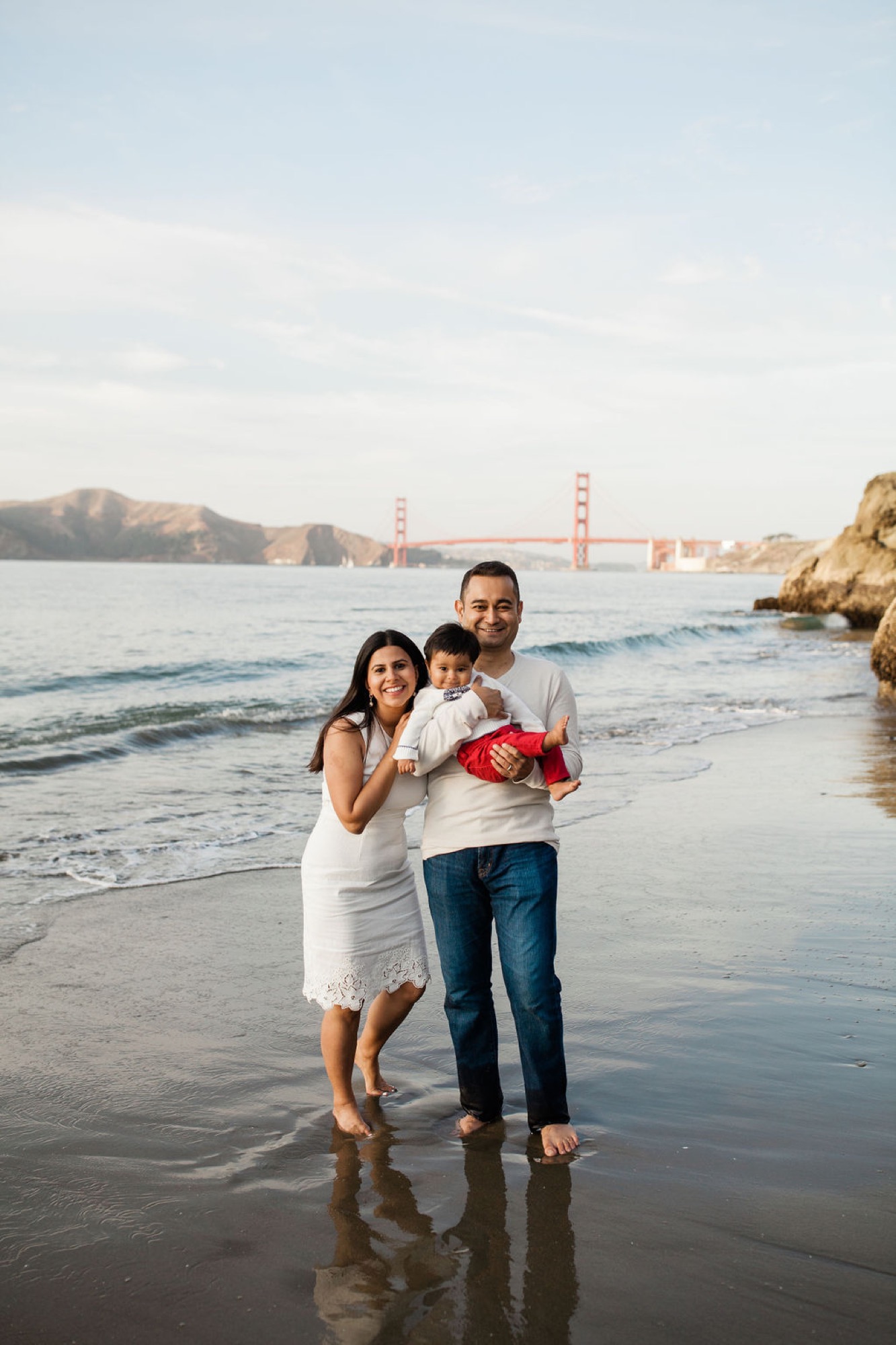 Pregnant mother and father hold their toddler son while standing in shallow water, the Golden Gate Bridge glowing behind them in golden afternoon light