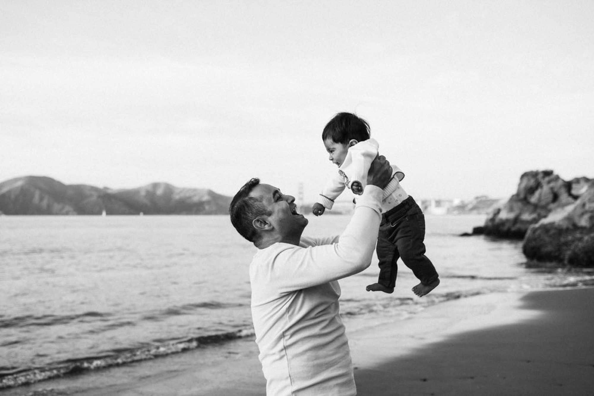 A father lifts his young son high against his chest, both smiling with joy as waves lap the sandy beach behind them
