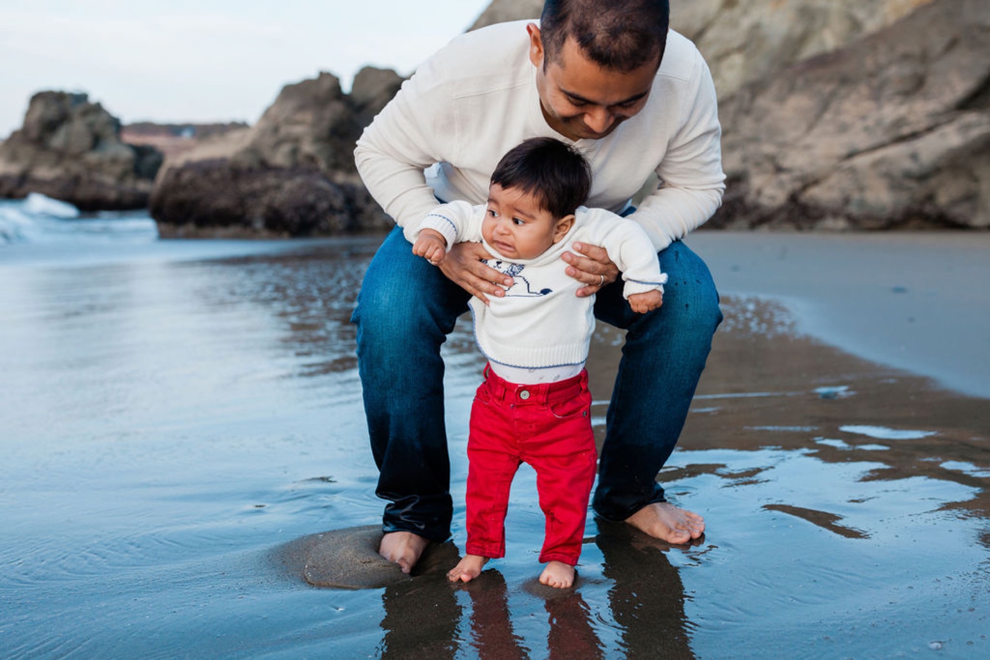 A father steadies his barefoot toddler in shallow tide pools, the child's uncertain expression reflecting newfound confidence in the water