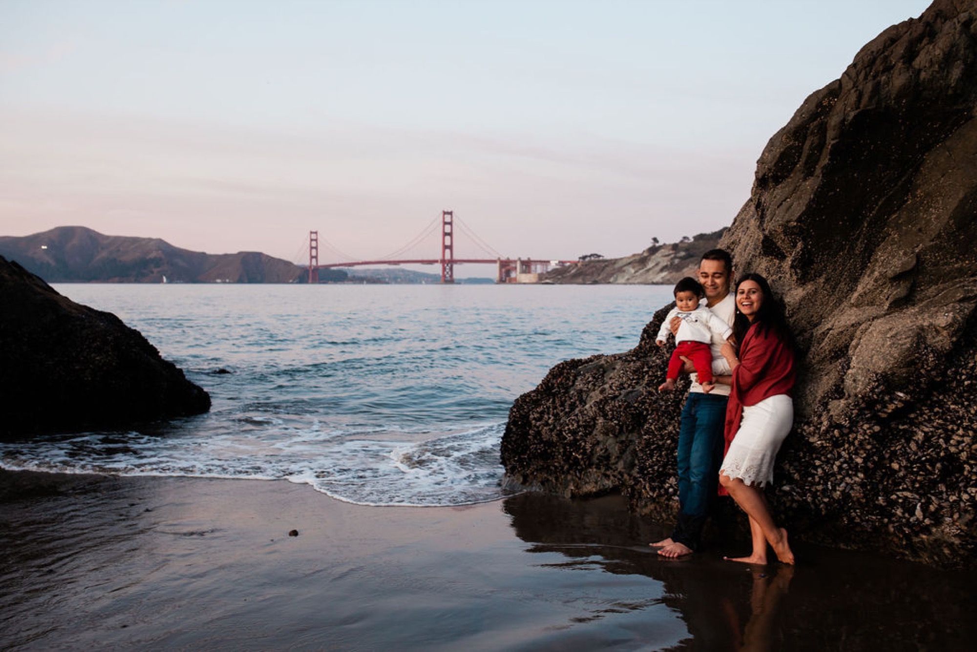 Family of three stands barefoot on rocky tide pools with the Golden Gate Bridge glowing softly in the evening light behind them