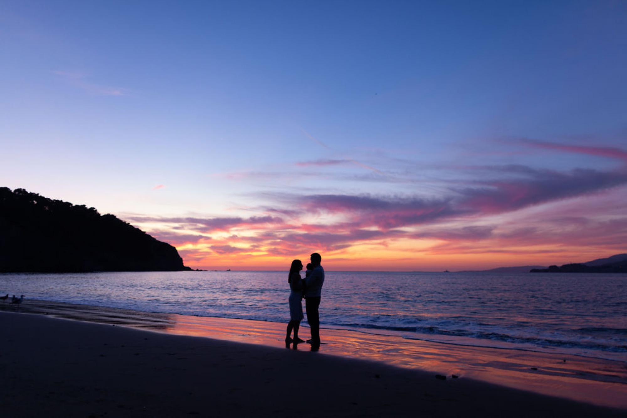 Two silhouetted figures embrace on the wet sand as golden sunset light reflects across the beach and dramatic clouds paint the sky