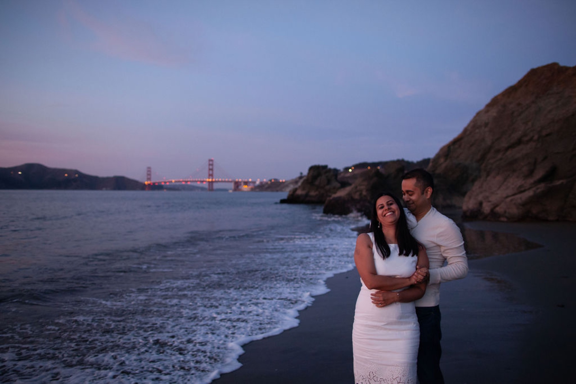 Couple embraces at twilight on China Beach with the illuminated Golden Gate Bridge glowing across the water behind them