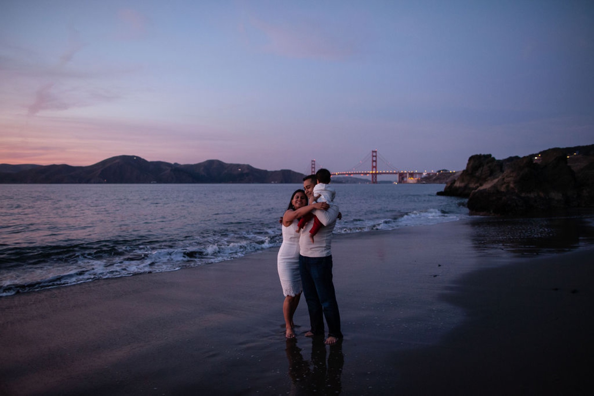 A man holds his young child while a woman embraces them both on the wet sand as the Golden Gate Bridge glows at twilight