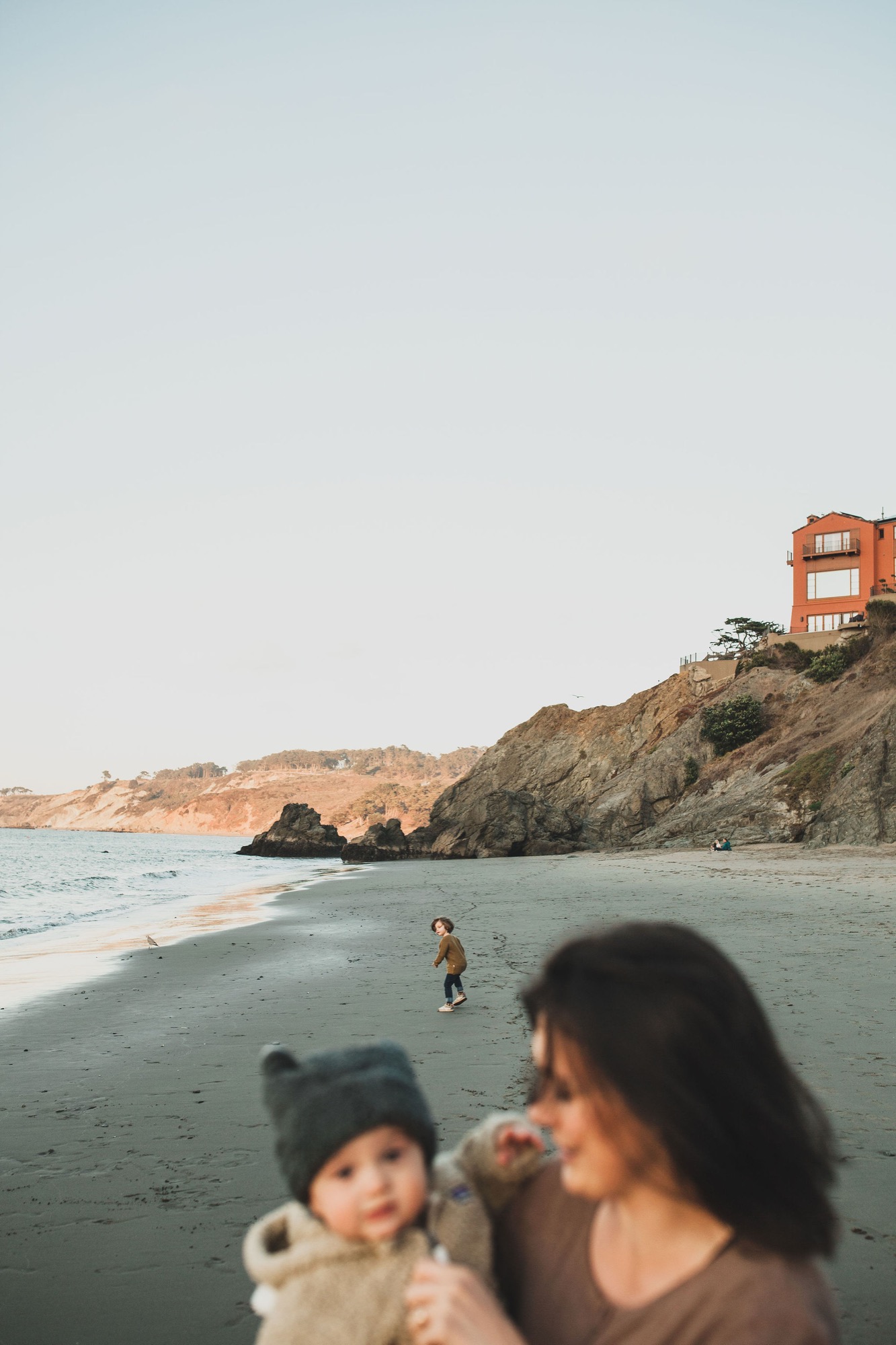 Woman holds toddler while watching child wade in shallow water beneath coastal cliffs and an orange house