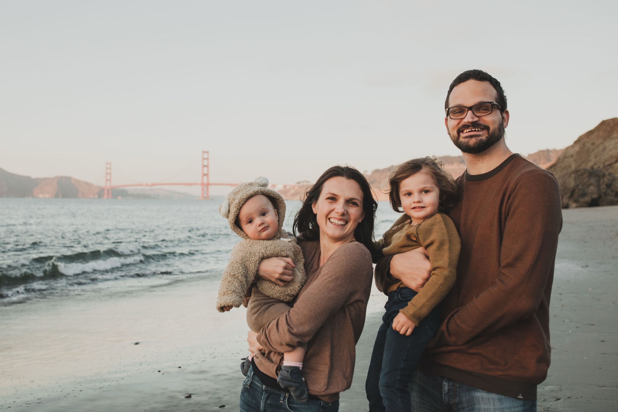 Family of four smiling at the camera on a beach with the Golden Gate Bridge visible in the misty background
