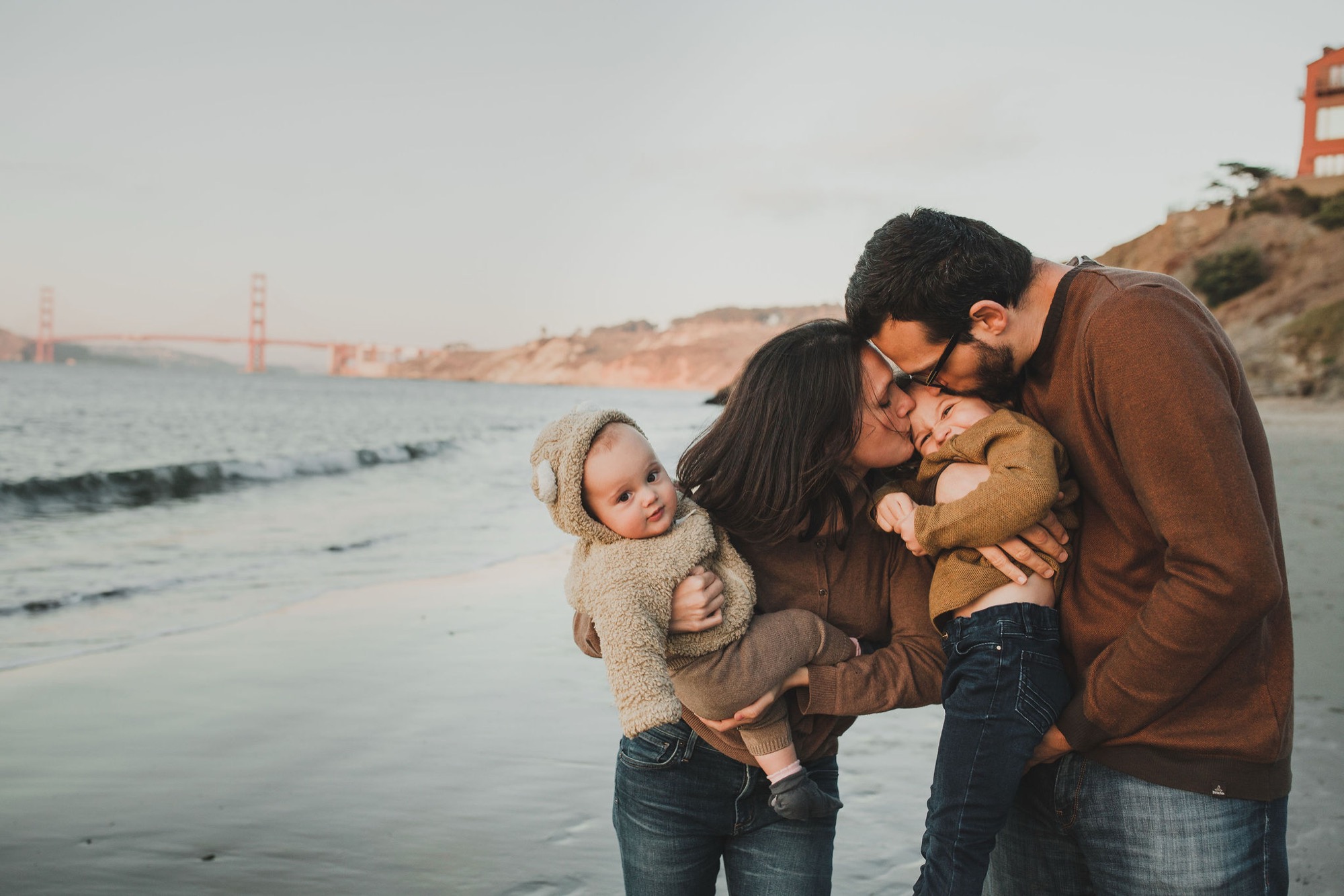 Parents kiss while holding their two young children on the beach at golden hour, with the Golden Gate Bridge visible across the water