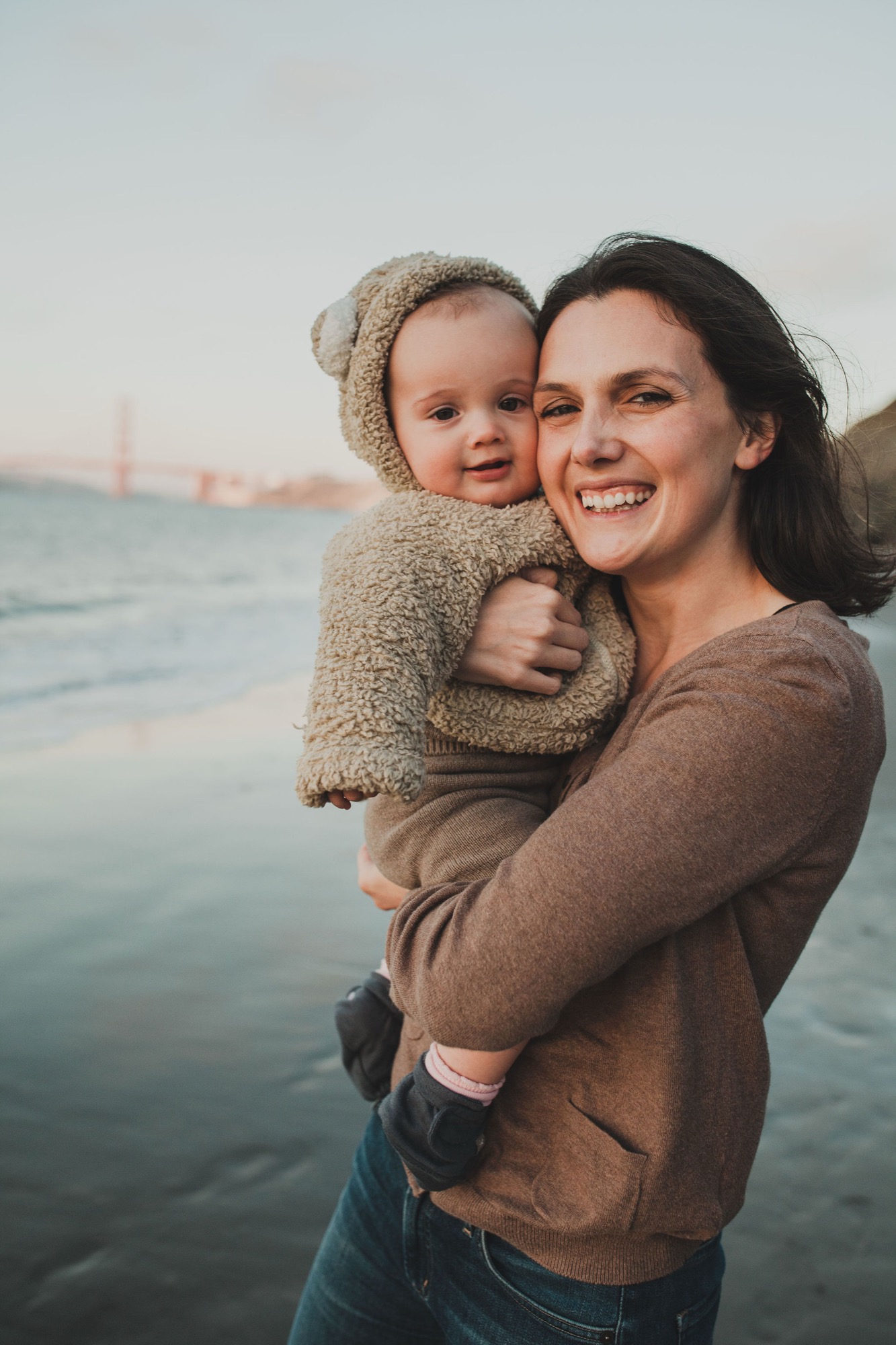 A smiling woman holds her toddler wrapped in a cozy knit blanket while standing on the beach at golden hour, with the Golden Gate Bridge visible in the misty distance