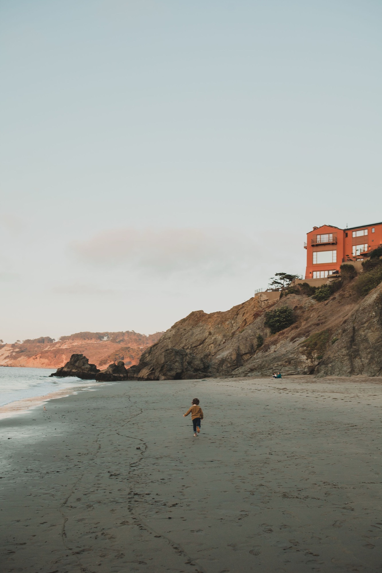 A solitary child wades into the cool Pacific waters while burnt-orange beach houses perch on the hillside above the sandy shore