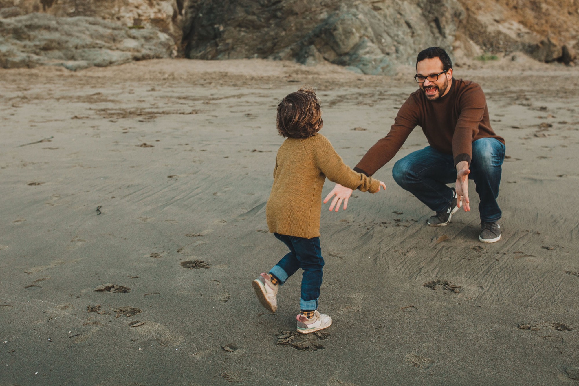 A father in glasses crouches to hold his toddler's hand on the wet sand, both moving toward the shoreline with rocky cliffs behind them