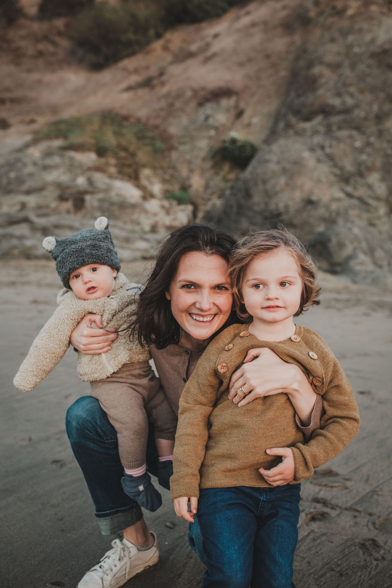 A smiling mother holds two young children on a sandy beach with eroded cliff faces rising behind them