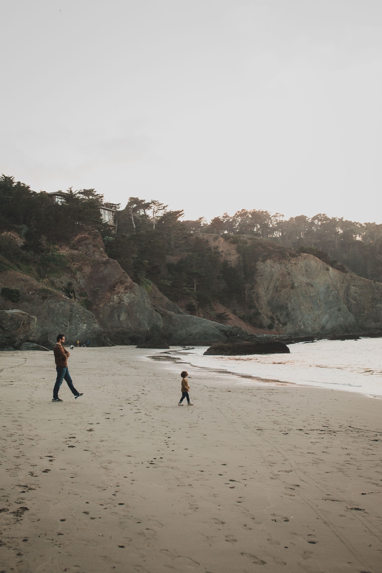 An adult and child walk across wet sand toward dramatic coastal cliffs under overcast skies, leaving footprints in their wake