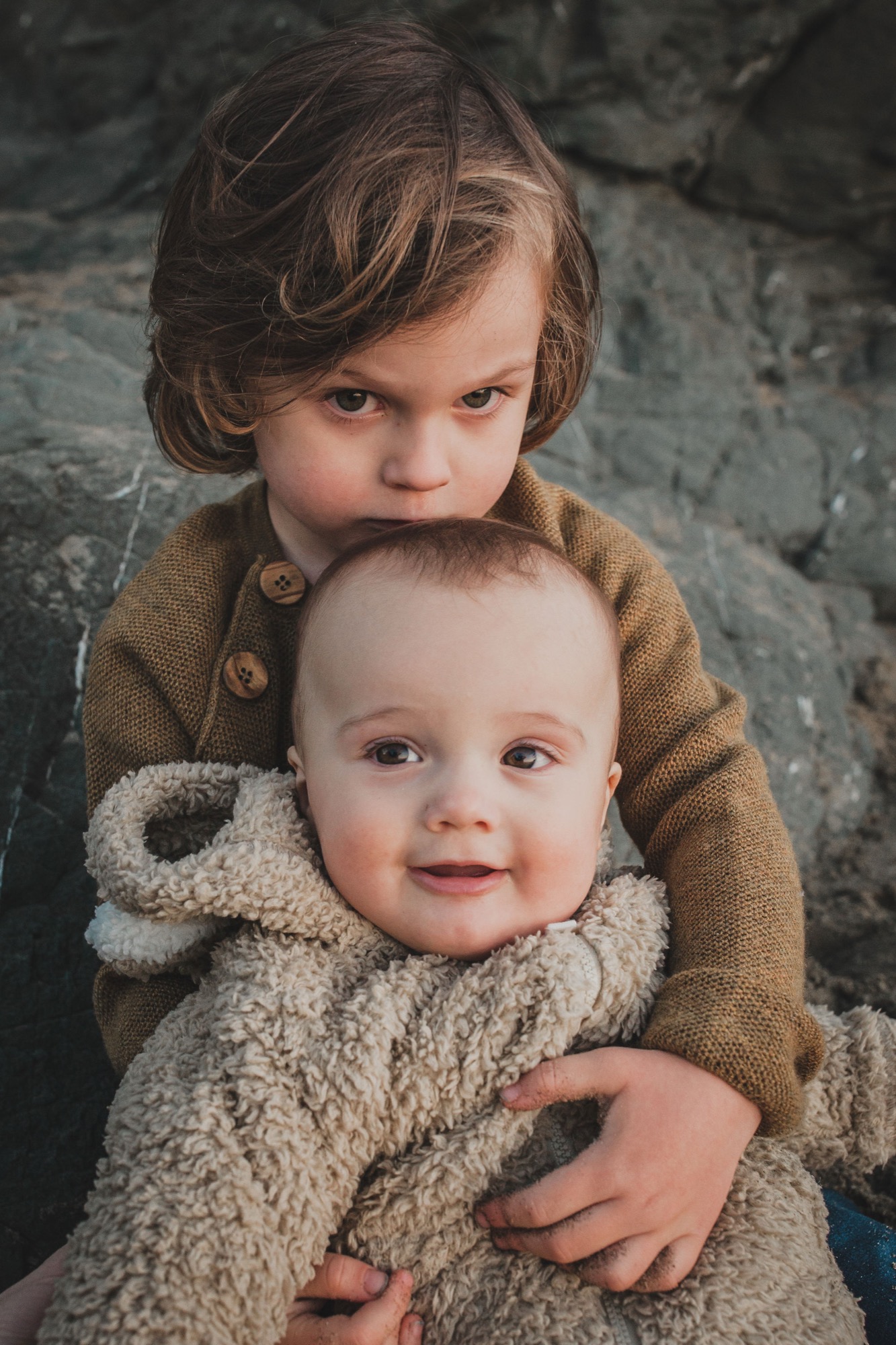 Older sister embraces younger toddler from behind against weathered stone, both gazing downward with gentle, contemplative expressions