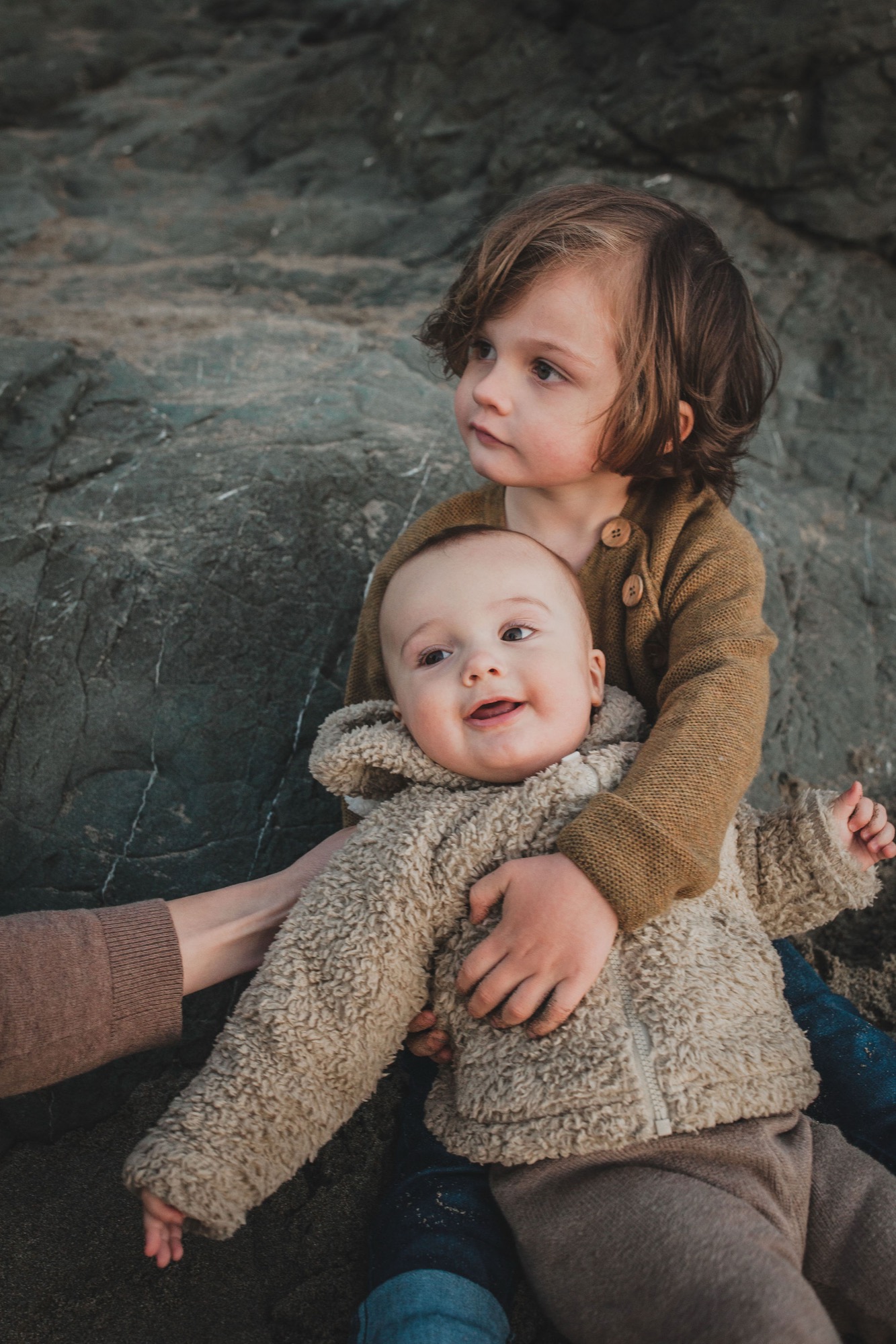 Two young children sit together on weathered rocks, the older girl gazing thoughtfully away while cradling her smiling baby sibling in a cream-colored sweater