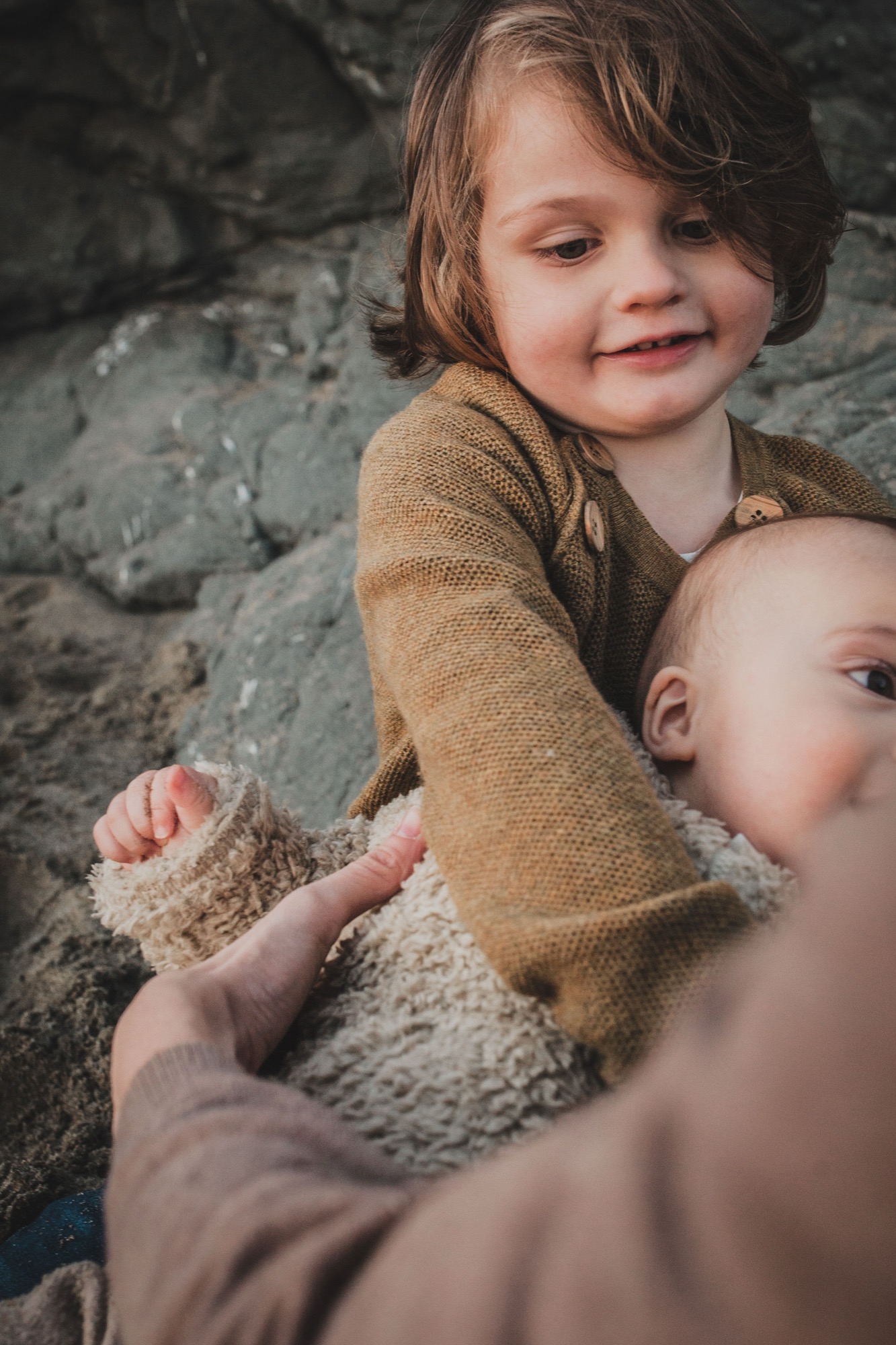 Two young children sit together on sandy ground, the older child in a mustard sweater gently holding the younger one's hand with genuine smiles