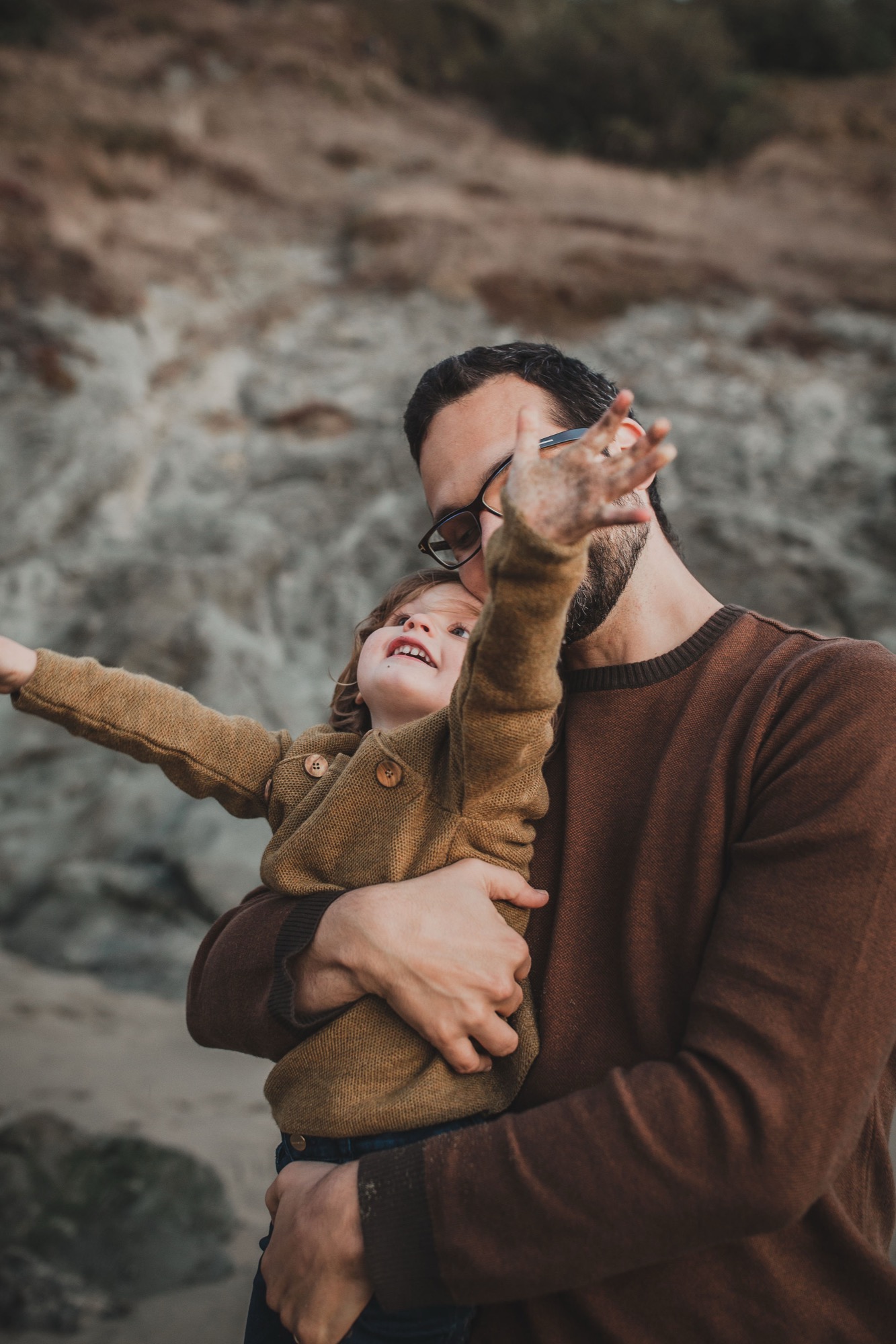 A father holds his young child aloft against weathered rock formations, both gazing upward with wonder and joy