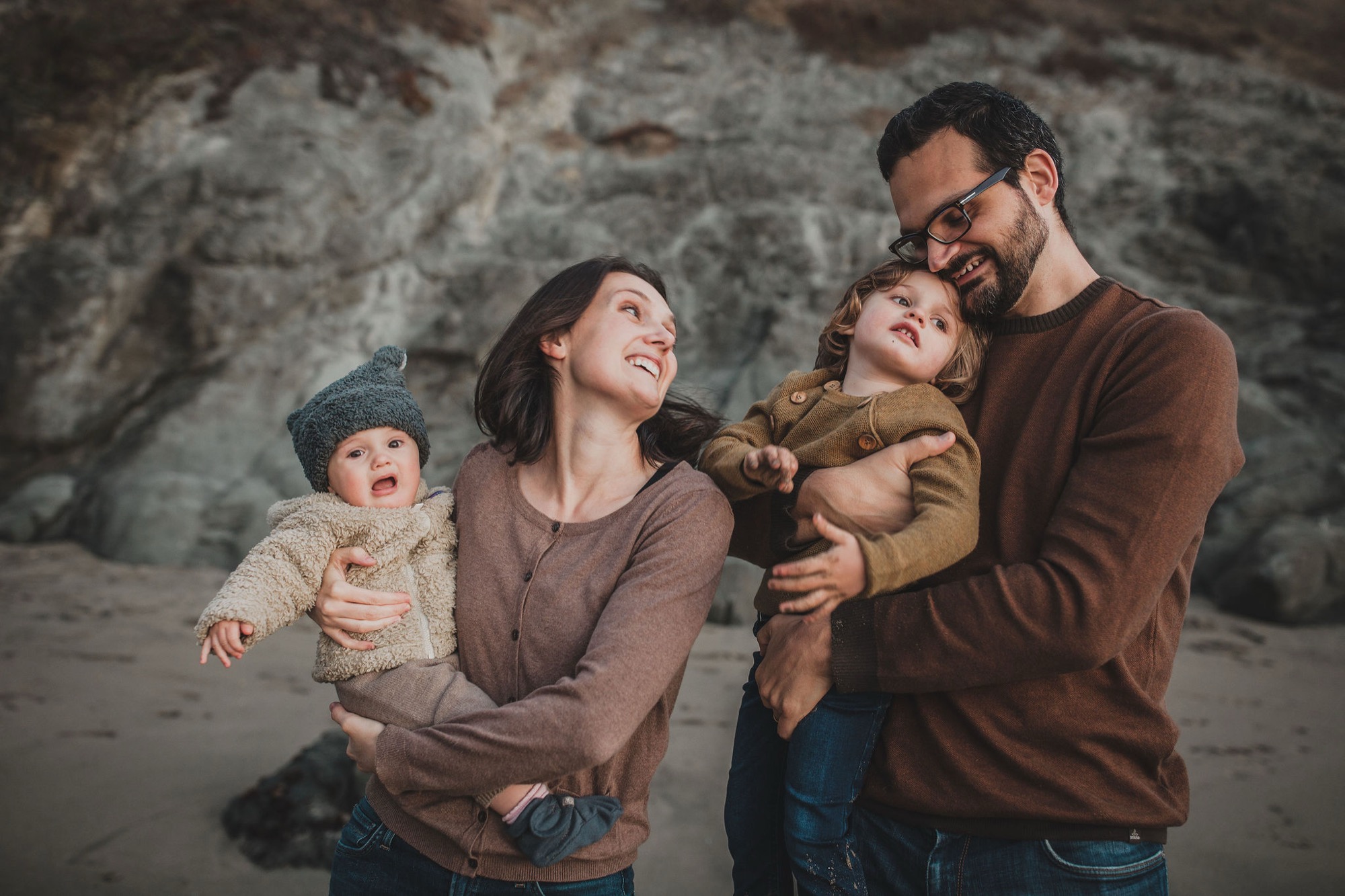A family of four stands together against weathered rock formations, with parents holding their two young children while gazing upward with genuine smiles