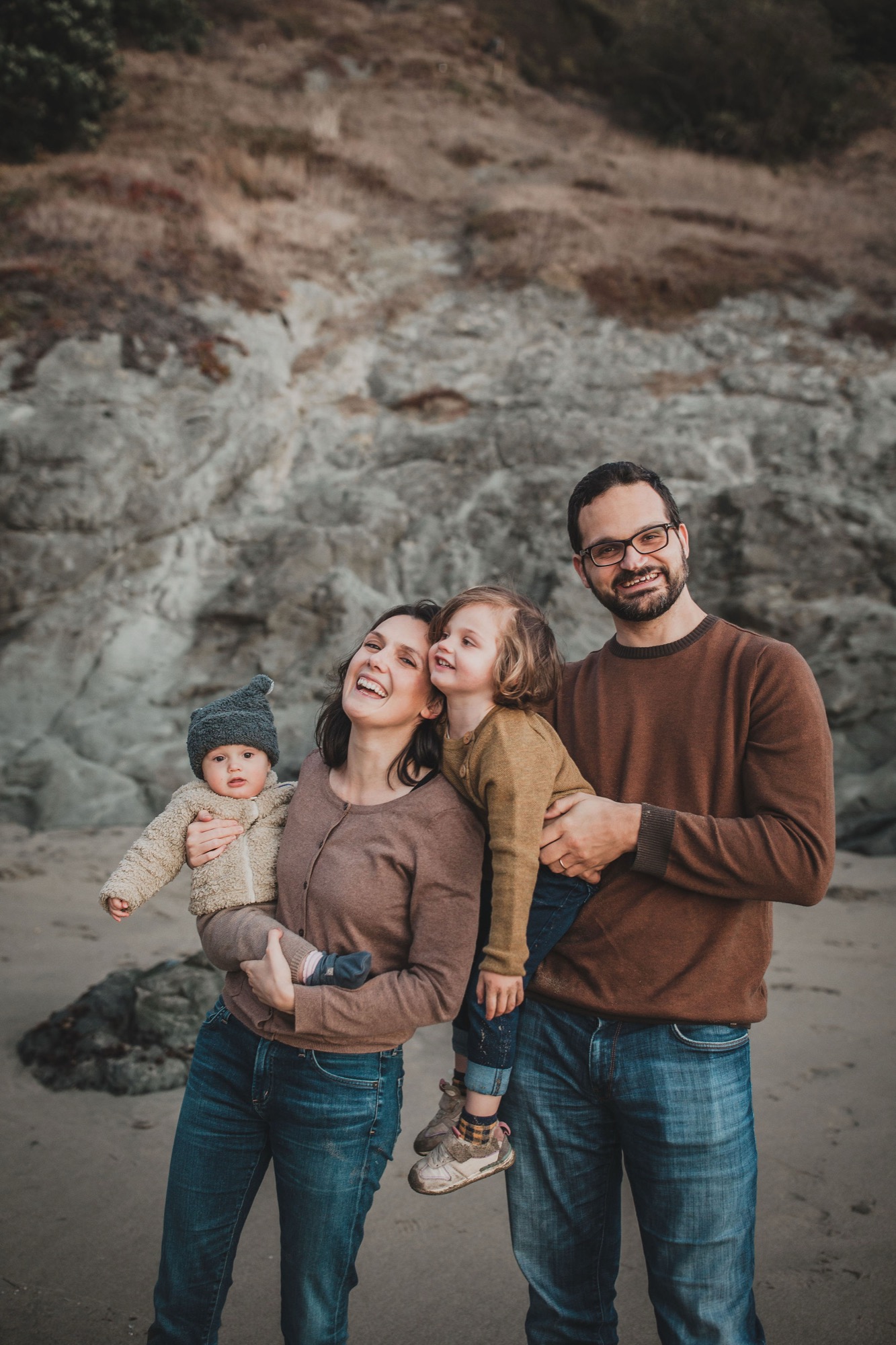 A family of four stands together on a beach with layered cliff formations behind them, all smiling warmly in coordinated earth-toned clothing