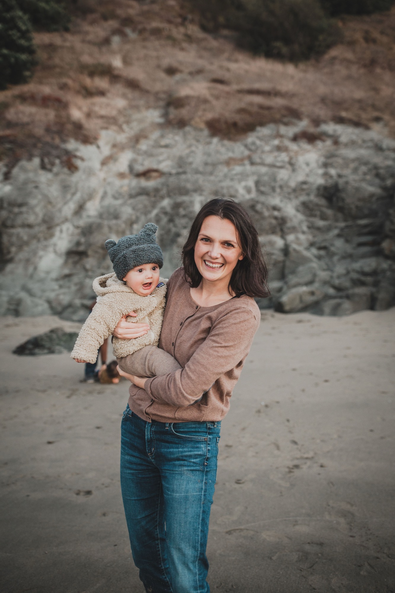 Woman in tan sweater holds smiling toddler in blue hat against dramatic weathered cliff face at beach