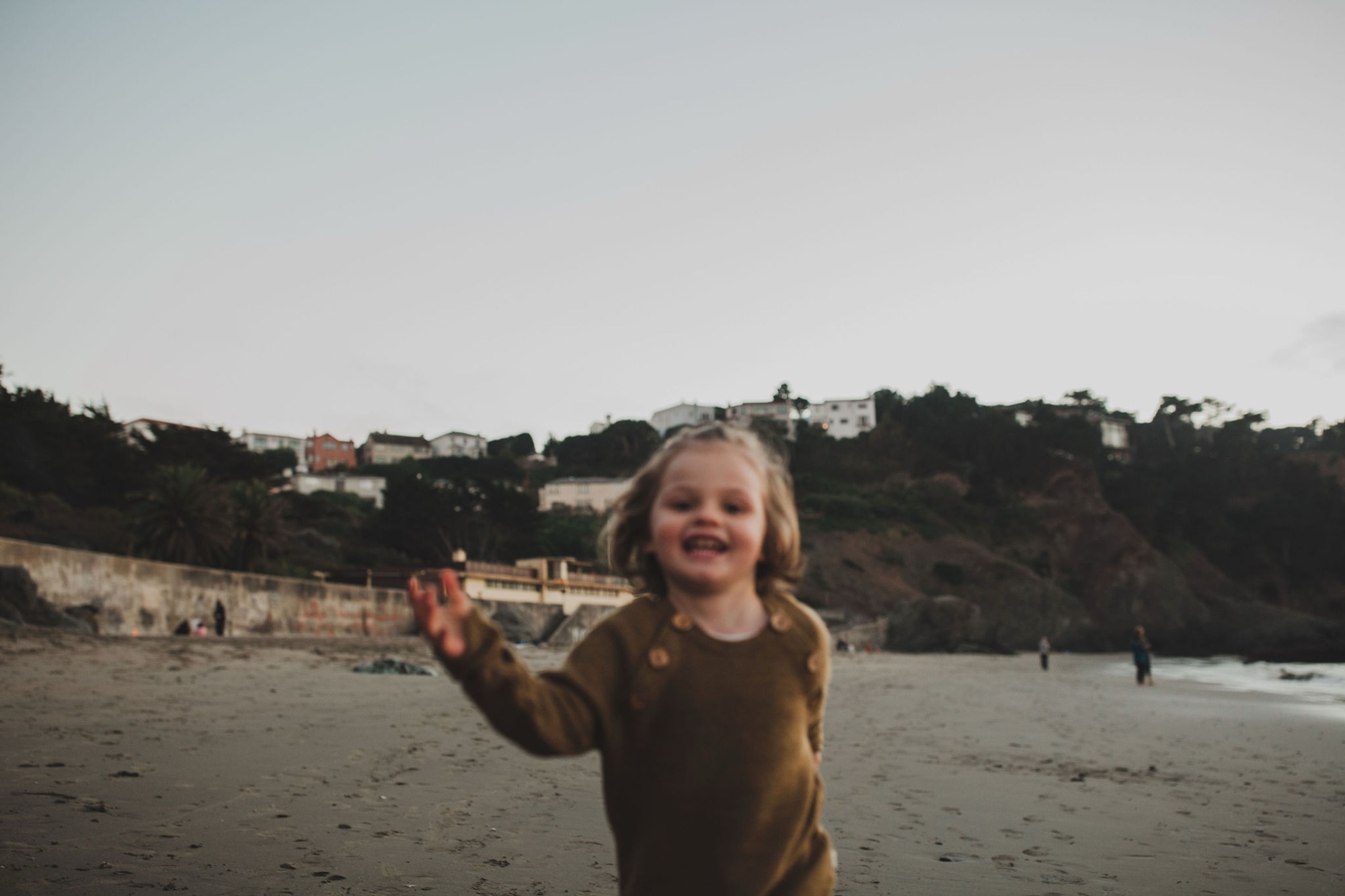 A blonde toddler in a brown sweater waves at the camera on a misty beach with cliffs rising behind her