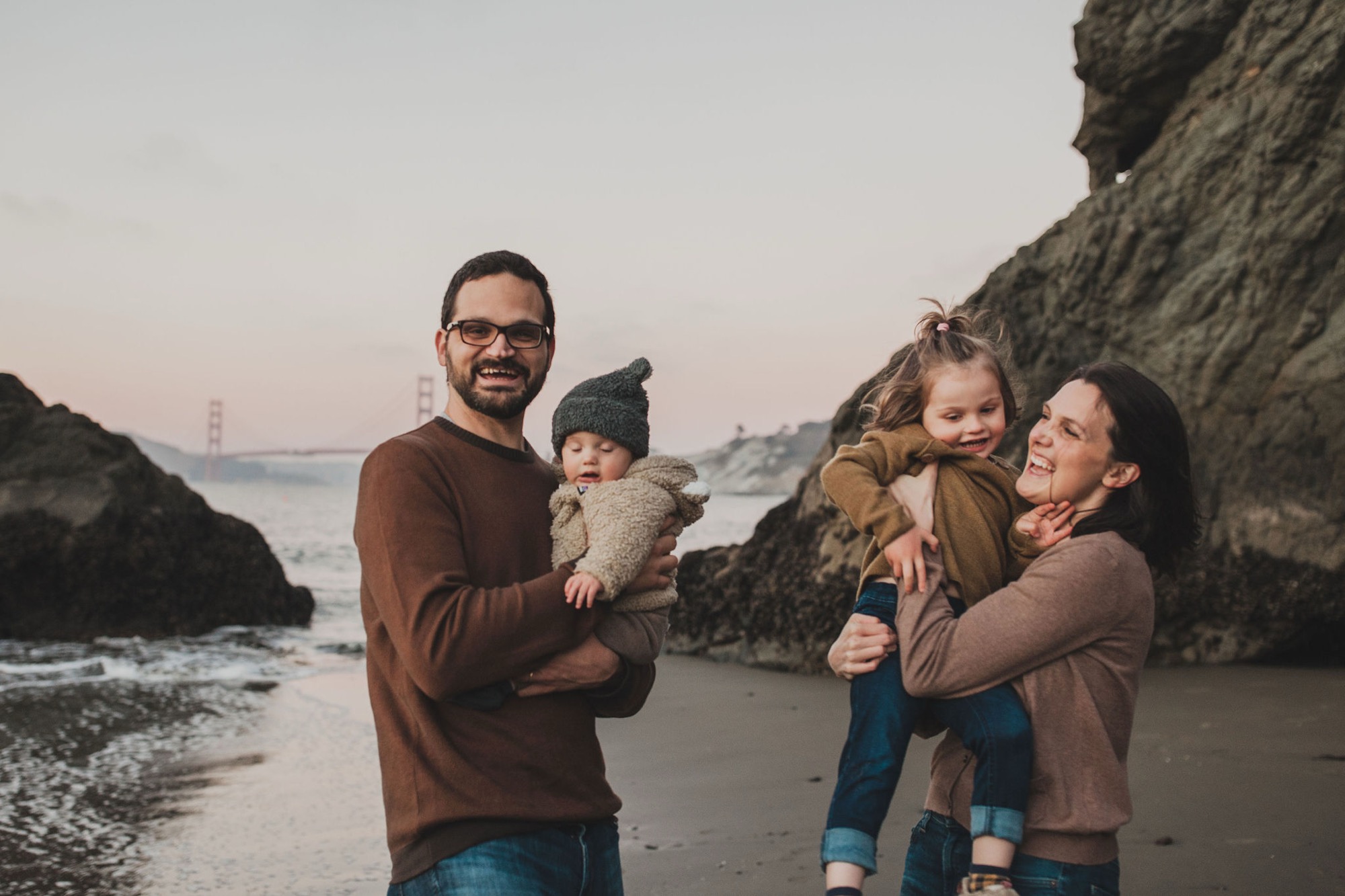 Two parents hold their young children on a rocky beach at sunset, all wearing coordinated brown tones while smiling at the camera