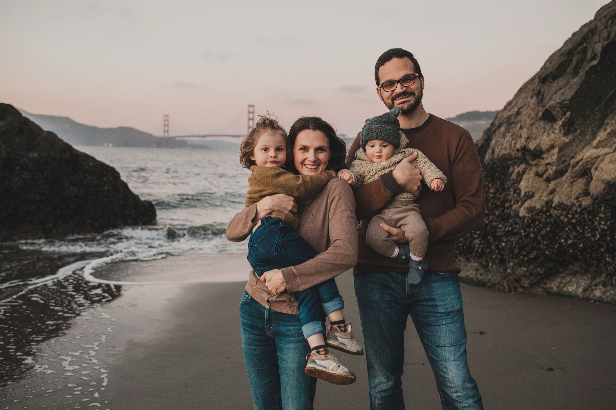 A bearded father and mother hold their two young children on wet sand at sunset, with the Golden Gate Bridge visible across the bay behind them